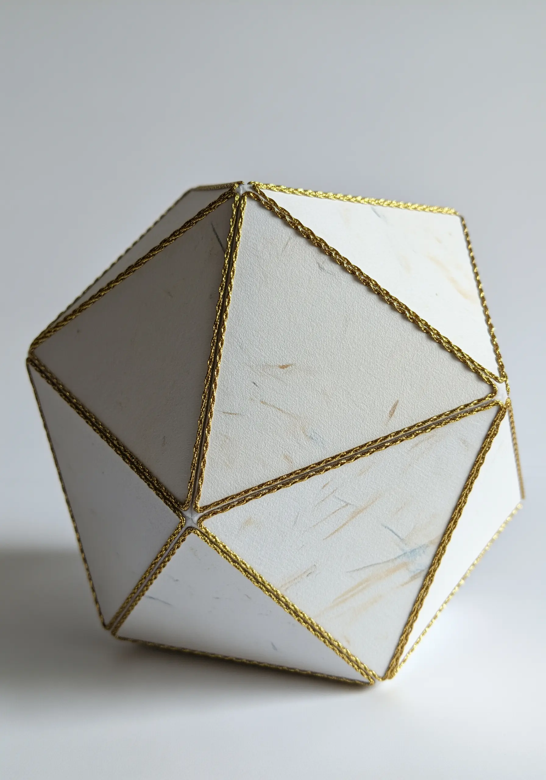 A white geometric paper icosahedron with its edges defined by a gold braided cord.
