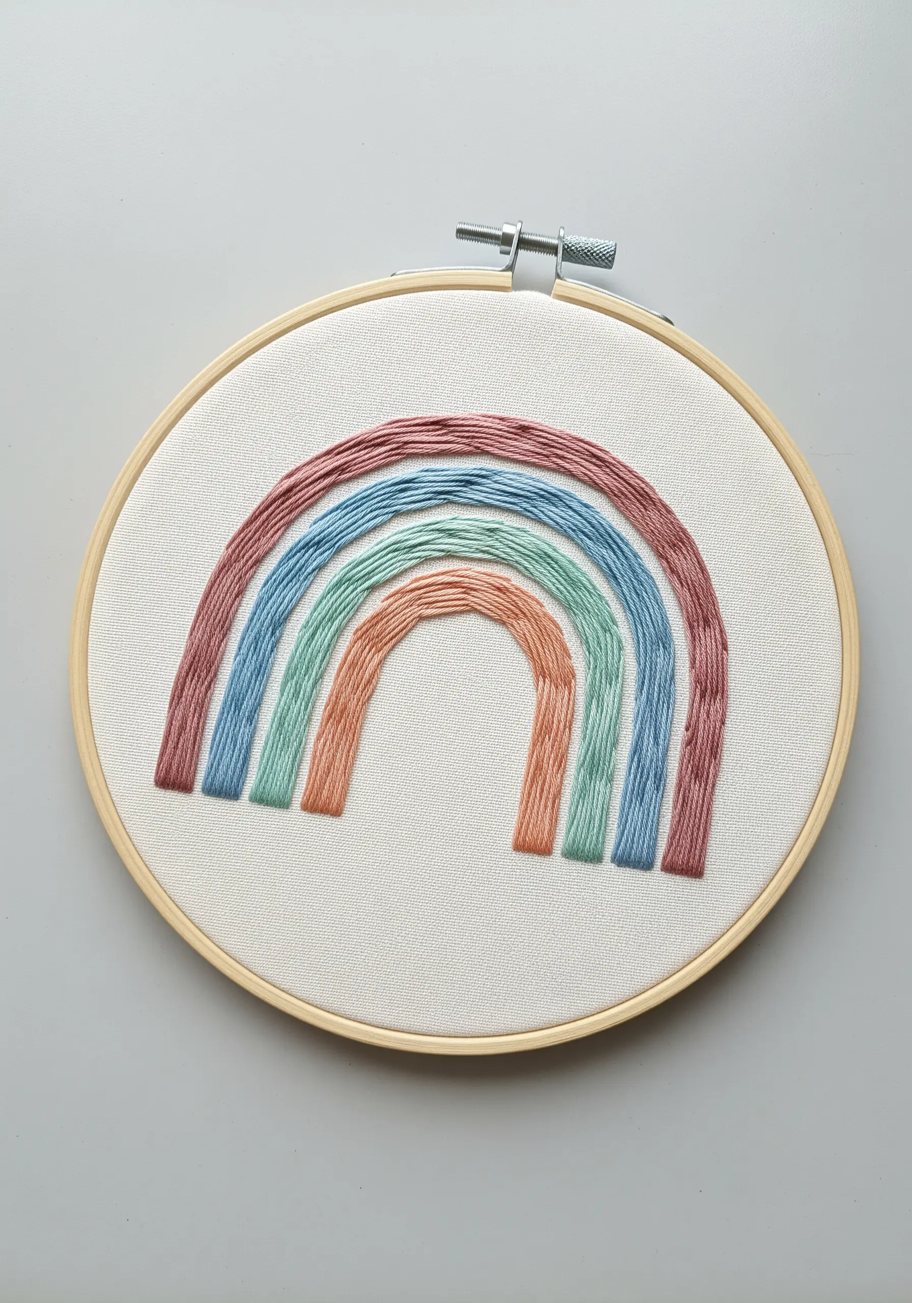 A modern rainbow embroidered in muted tones of dusty rose, blue, green, and terracotta.