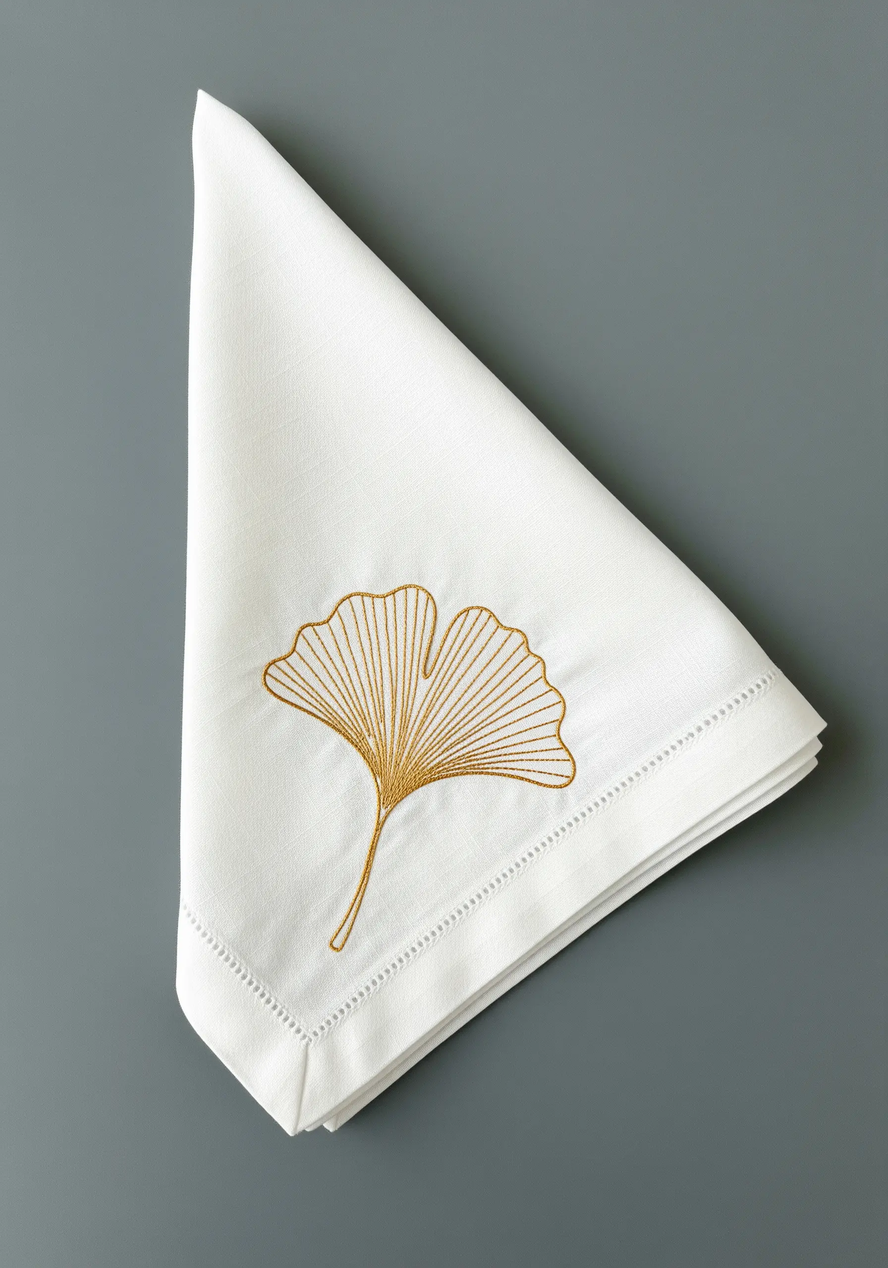 A golden ginkgo leaf embroidered on the corner of a folded white linen napkin.