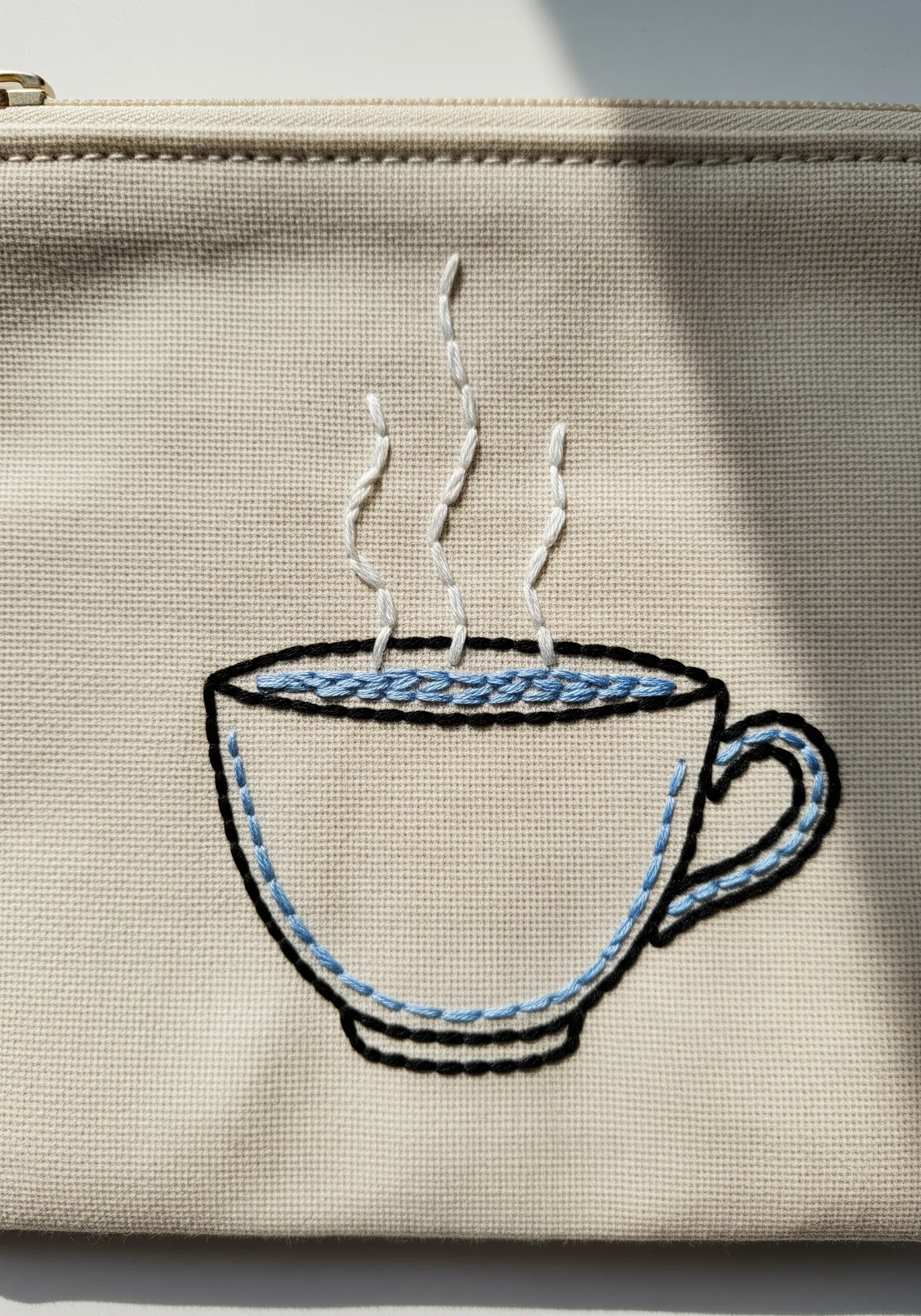 A line art embroidery of a coffee cup with blue accents, stitched on a textured pouch.
