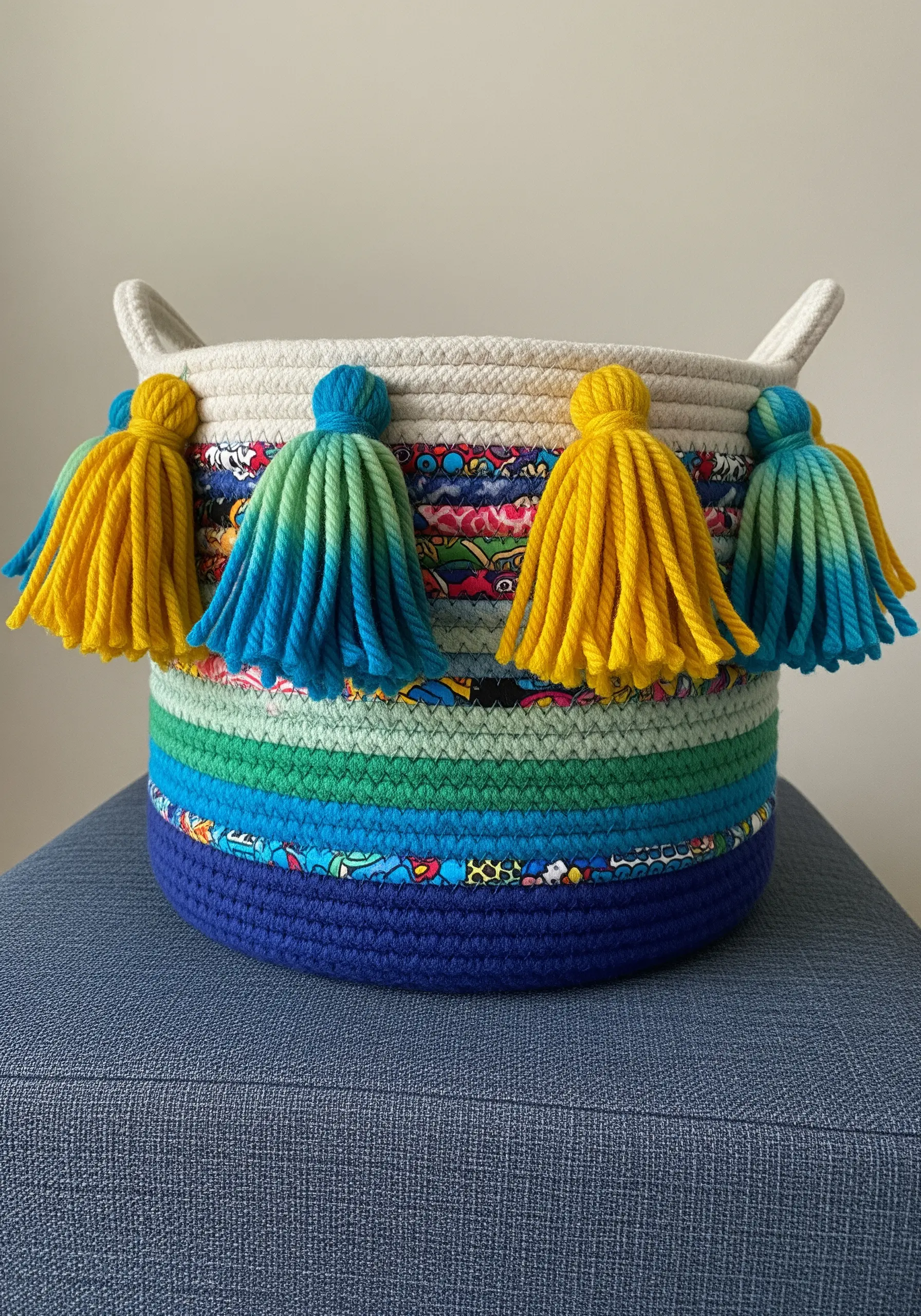 A coiled rope basket decorated with colorful fabric strips and bright yellow and blue tassels.