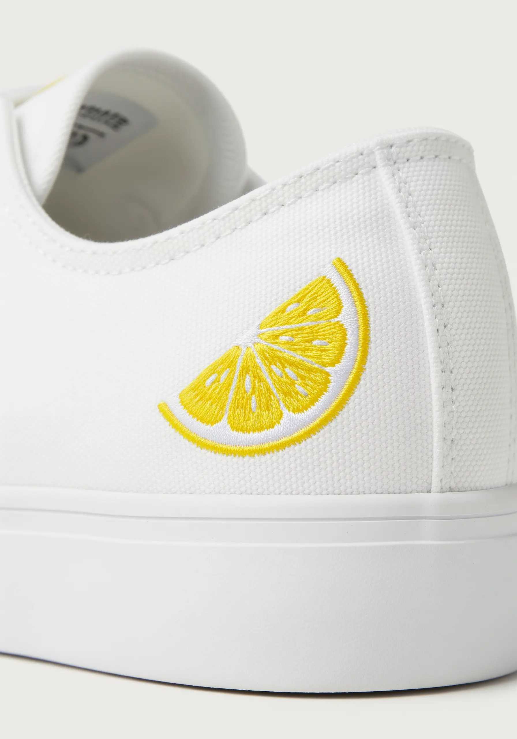 A bright yellow lemon slice embroidered on the heel of a white canvas sneaker.
