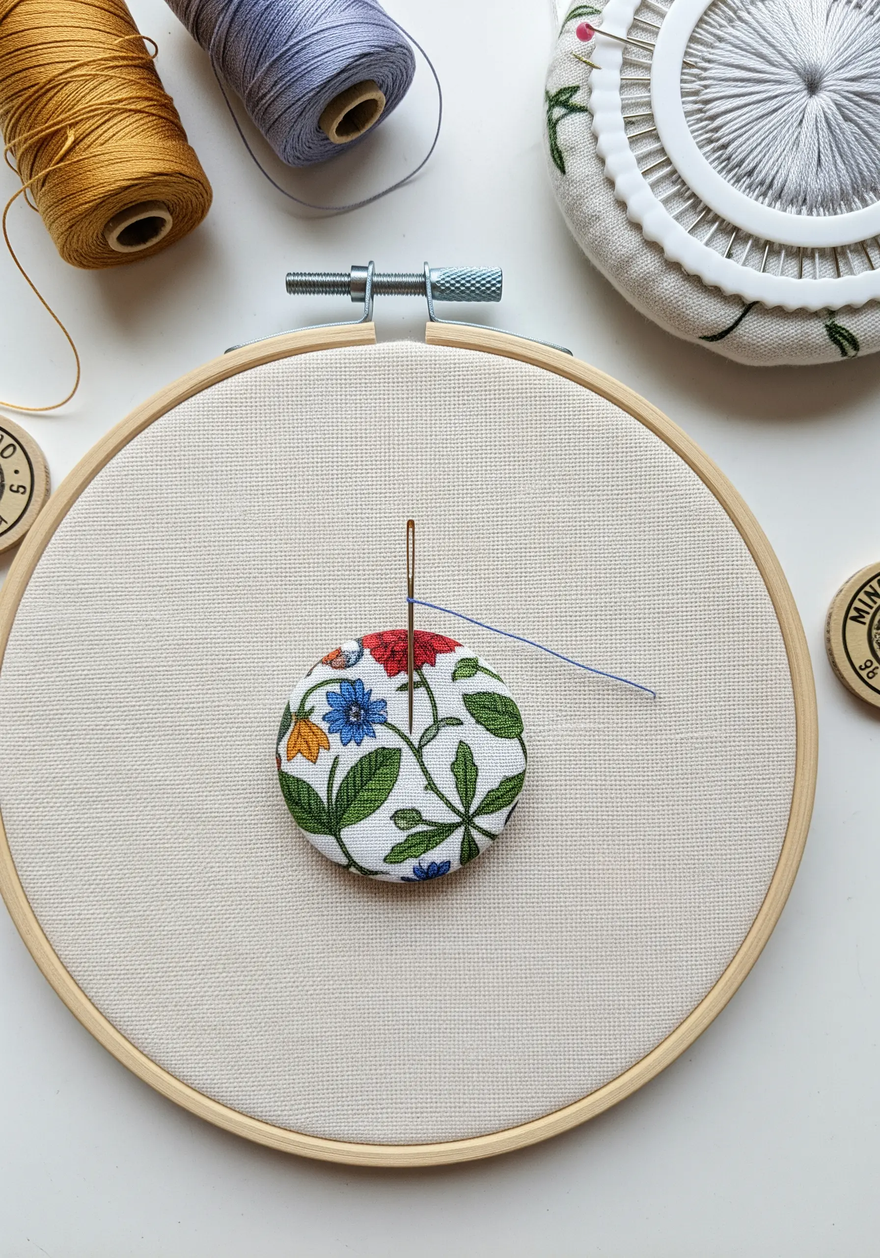 A fabric-covered button with a small floral motif being embroidered in a hoop.