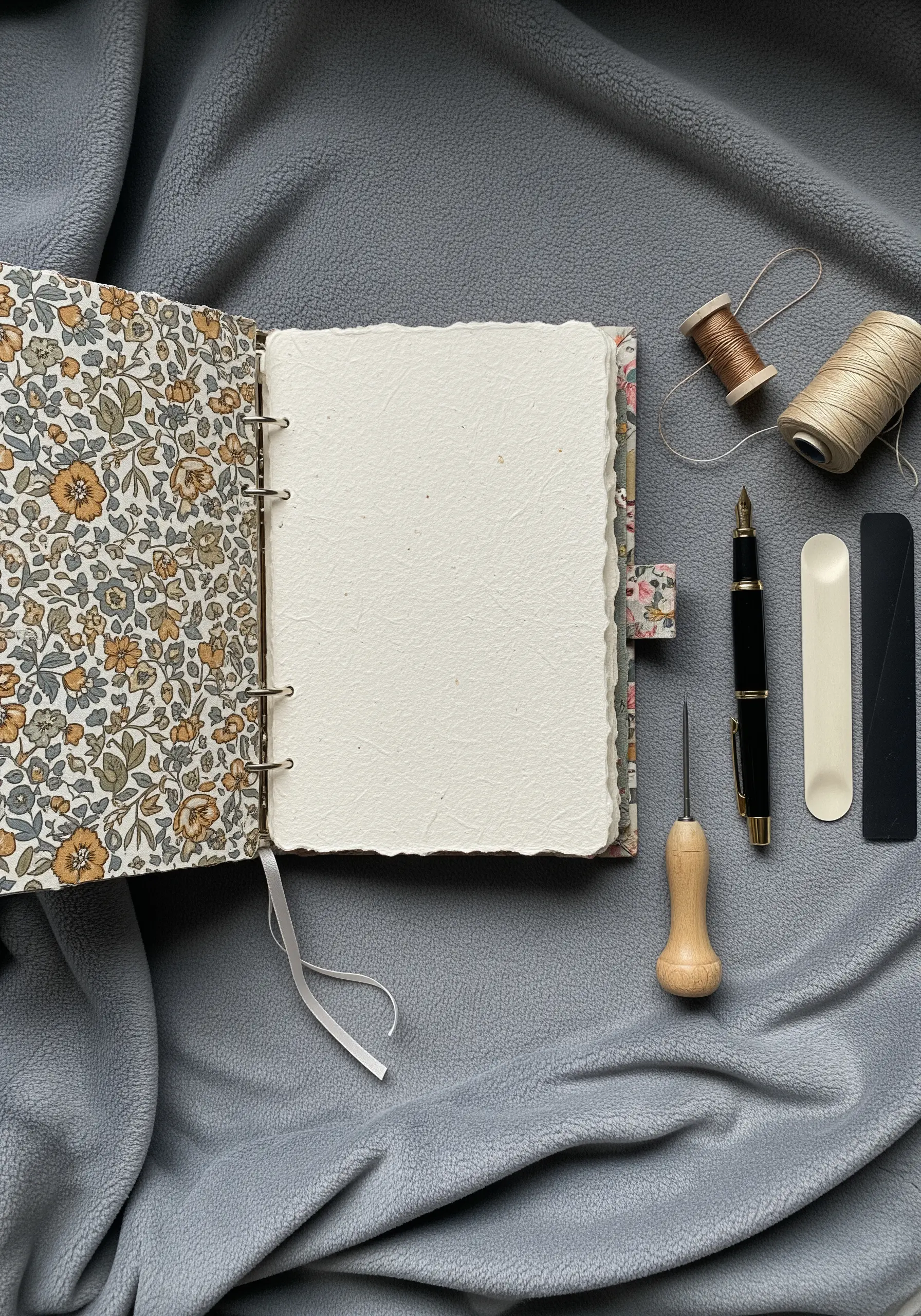 An open handmade journal with a floral fabric cover, next to bookbinding tools and thread.