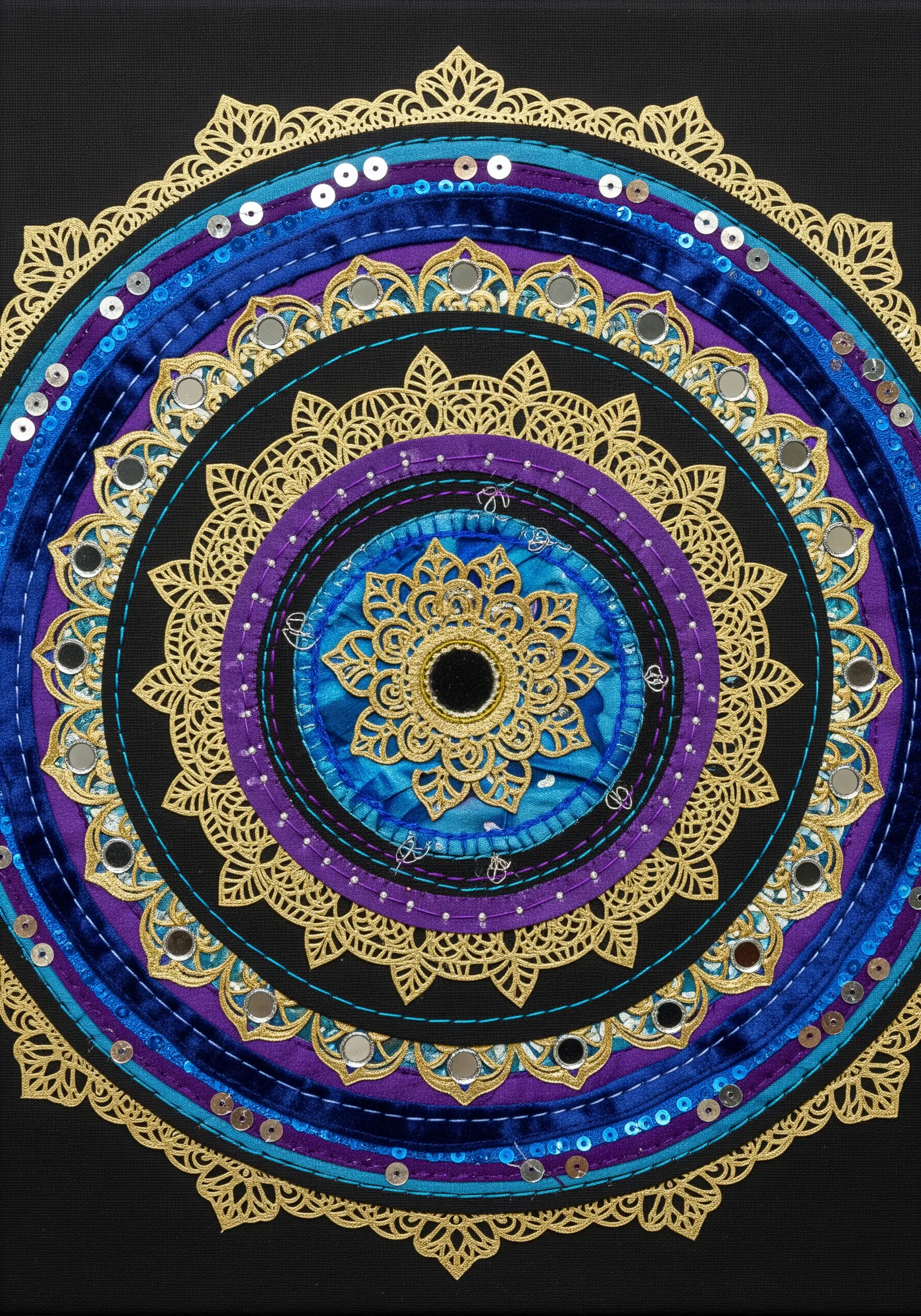 A layered mandala collage with a gold paper cutout over blue fabric, accented with sequins