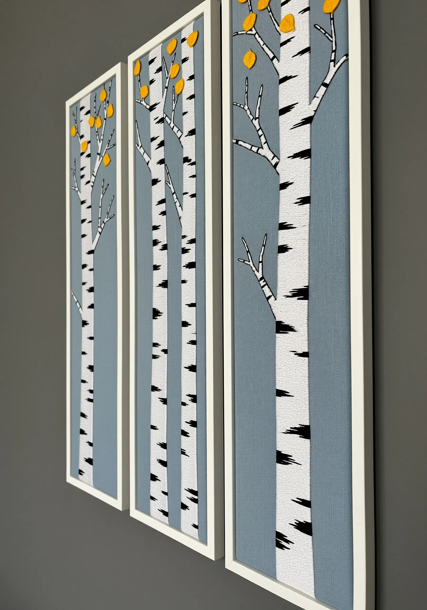 A triptych of framed embroidery showing minimalist birch trees against a dusty blue background.