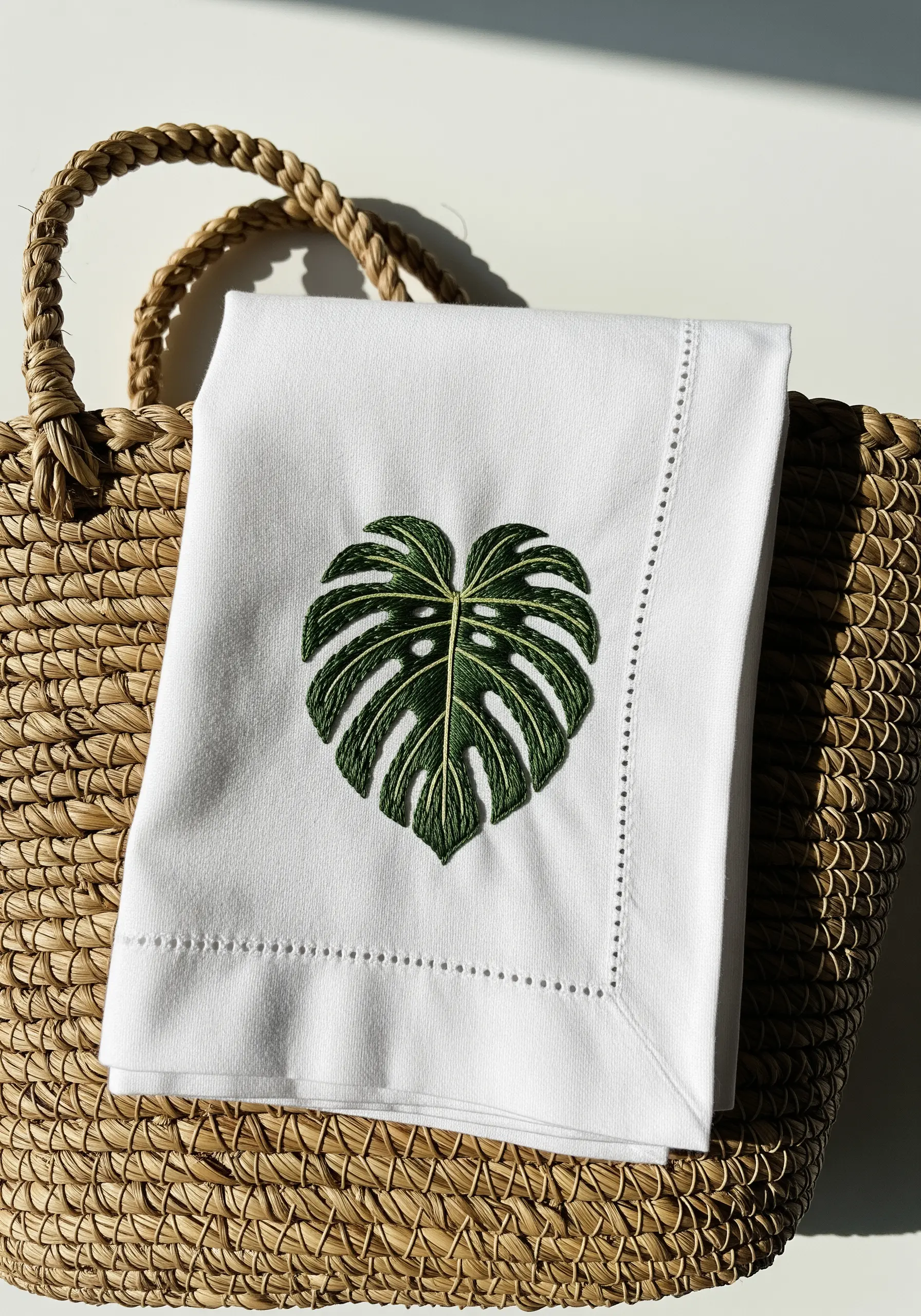 A single, deep green embroidered monstera leaf on a classic white hemstitched napkin.