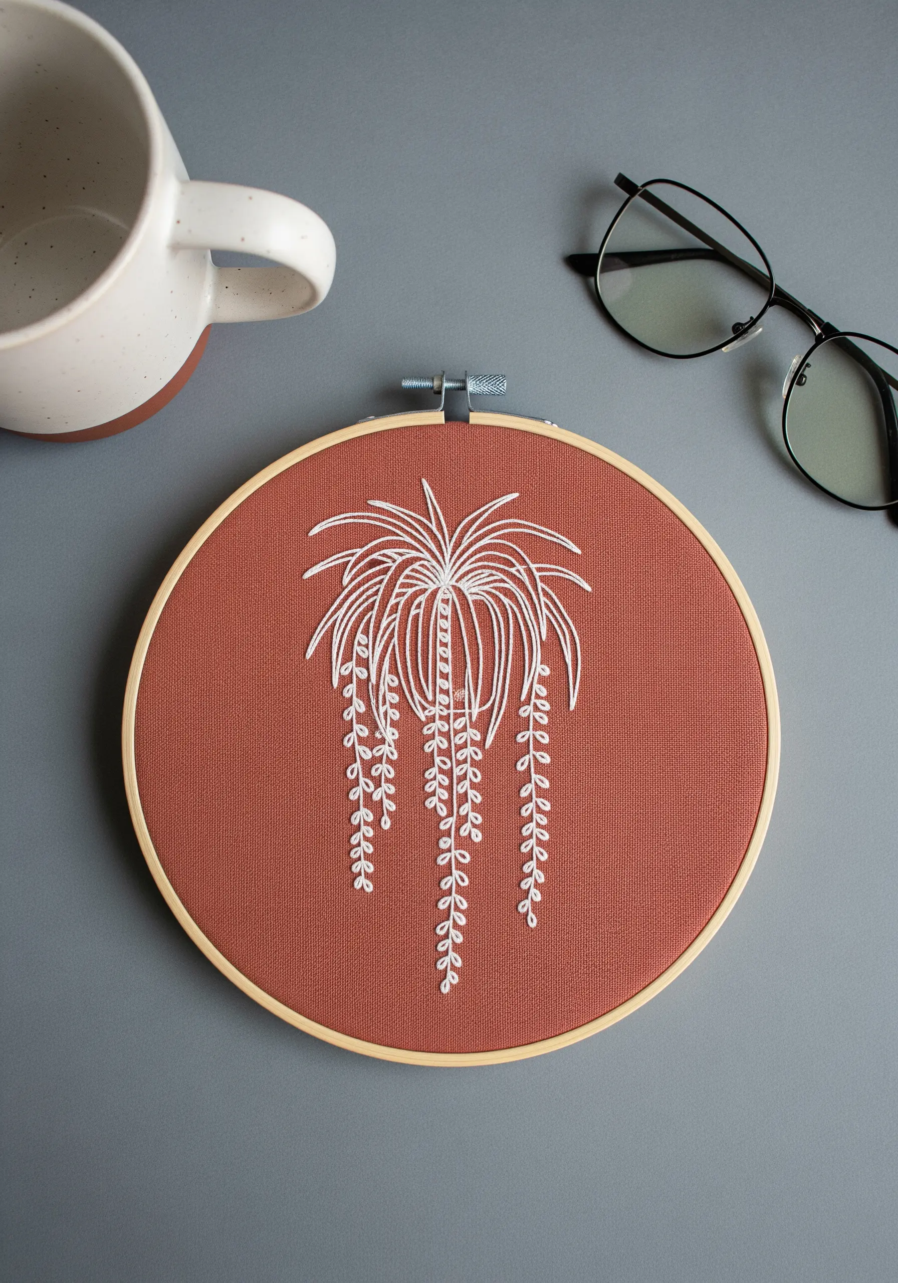 Minimalist line art of a trailing plant stitched in white on terracotta fabric.