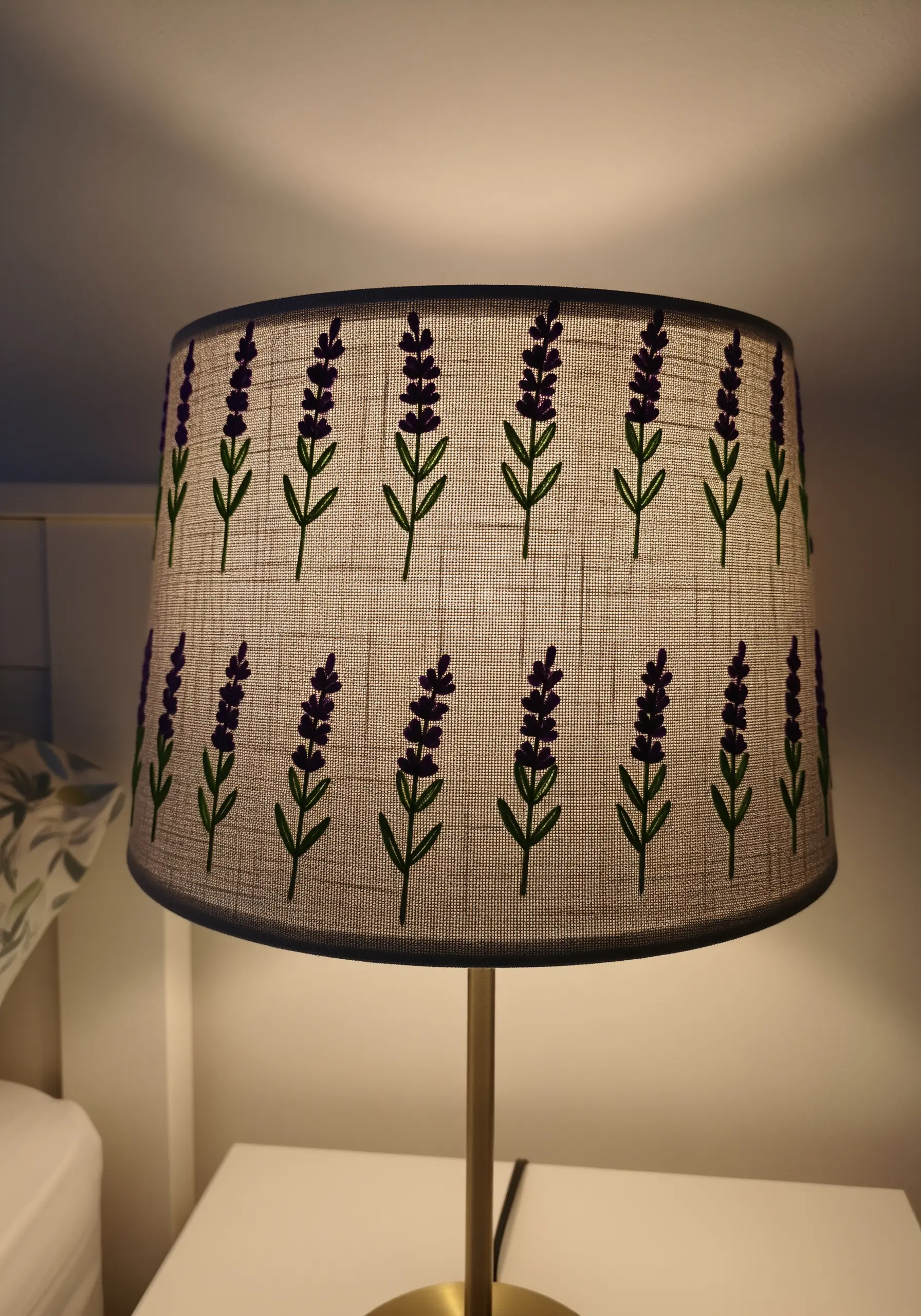 A fabric lampshade illuminated from within, showing an embroidered pattern of lavender sprigs.