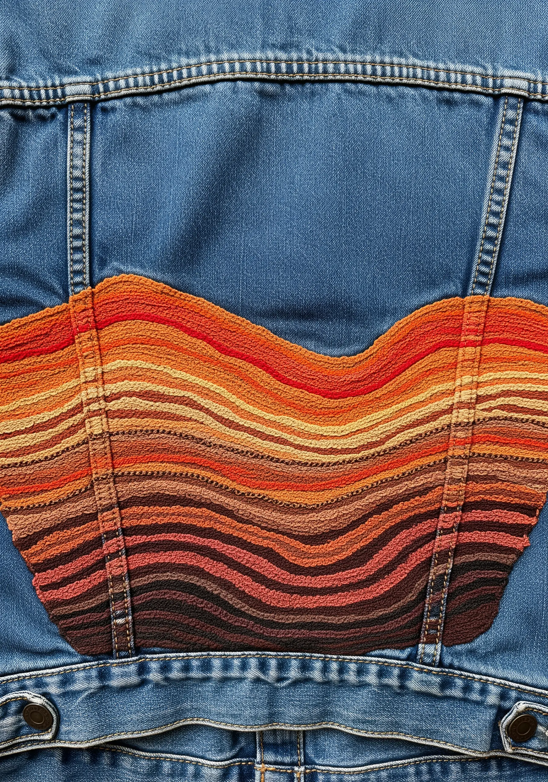 An abstract wavy shape on a denim jacket filled with a woven-textured stitch in earthy orange tones.