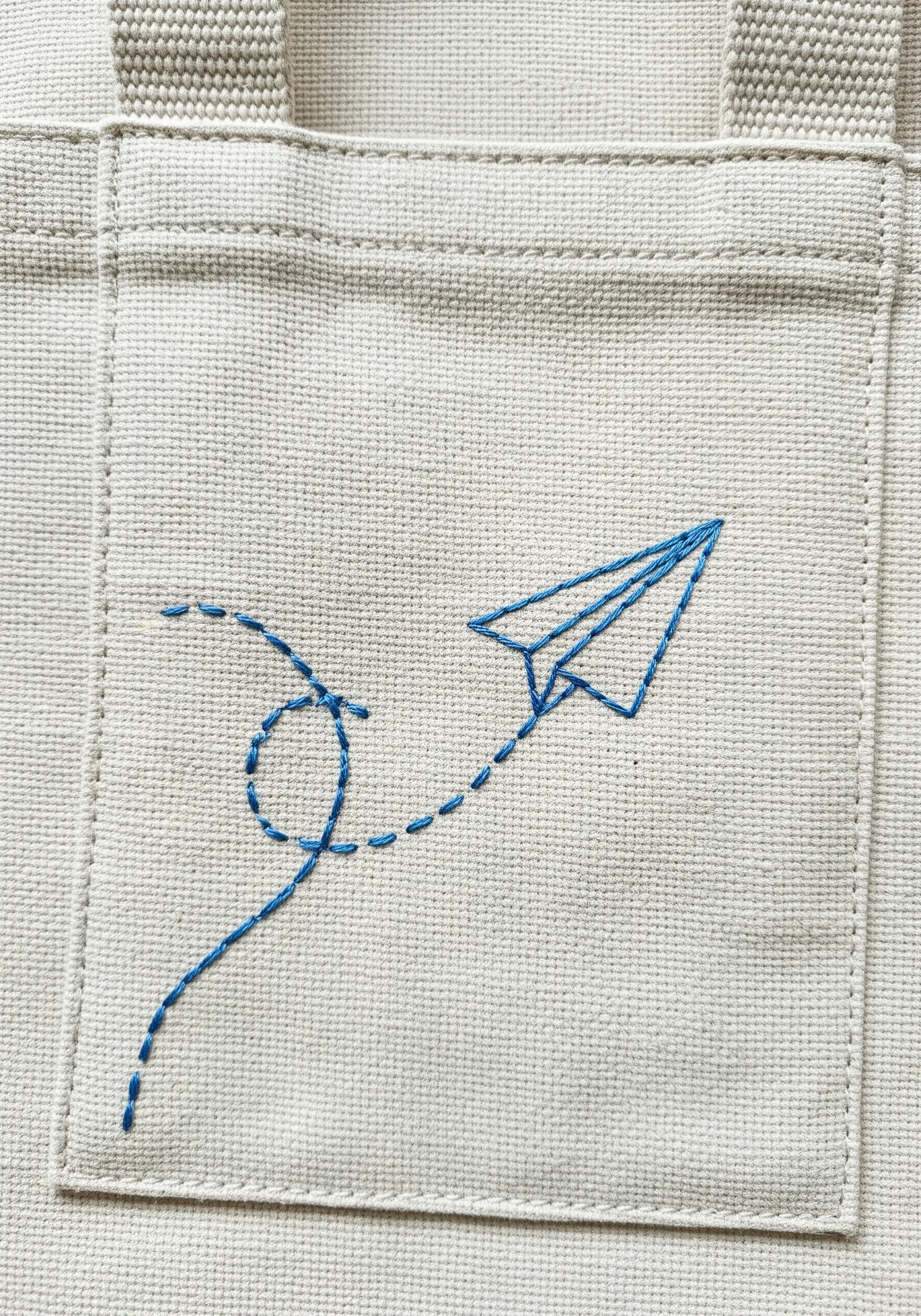 Blue paper airplane embroidered on a pocket with a dashed line trail behind it.