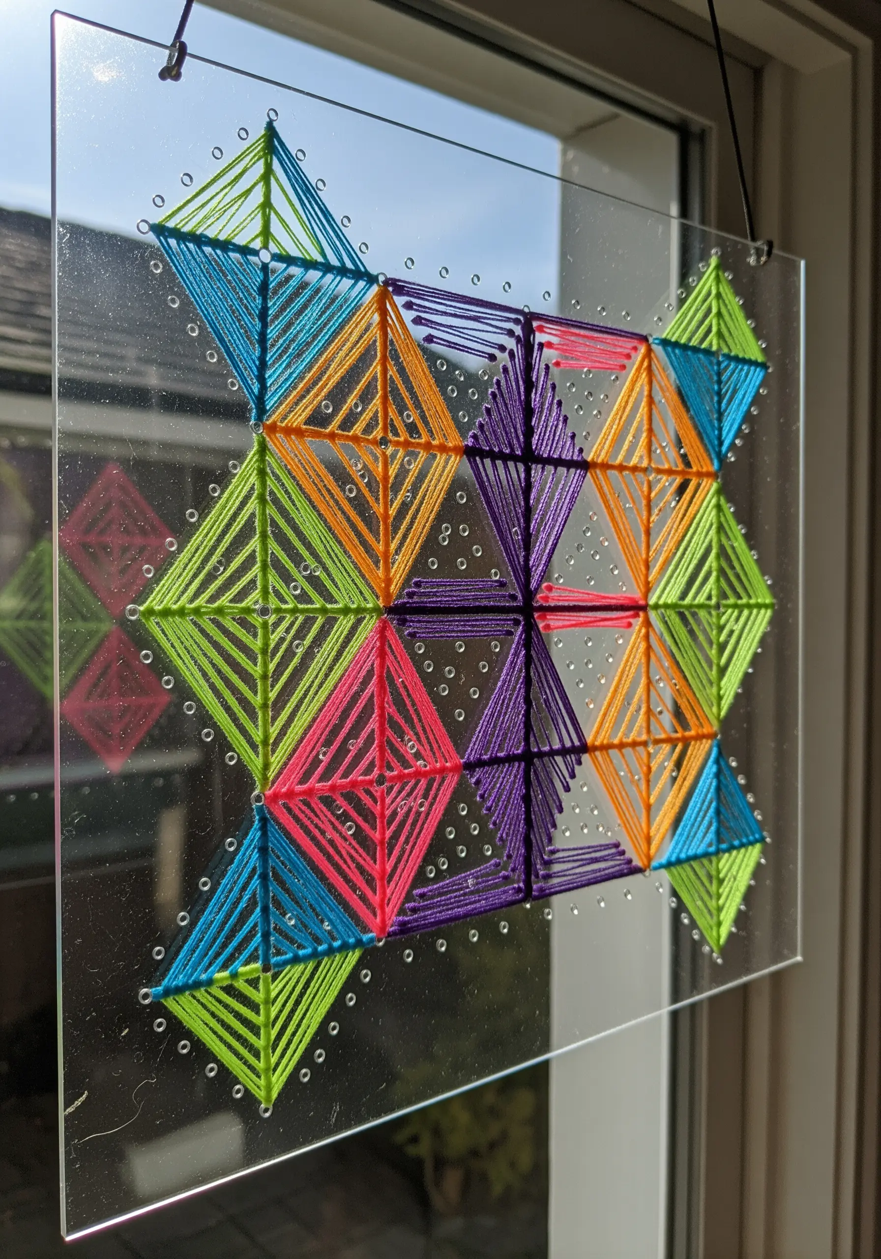Geometric string art made by weaving colorful thread through holes in a clear acrylic panel.