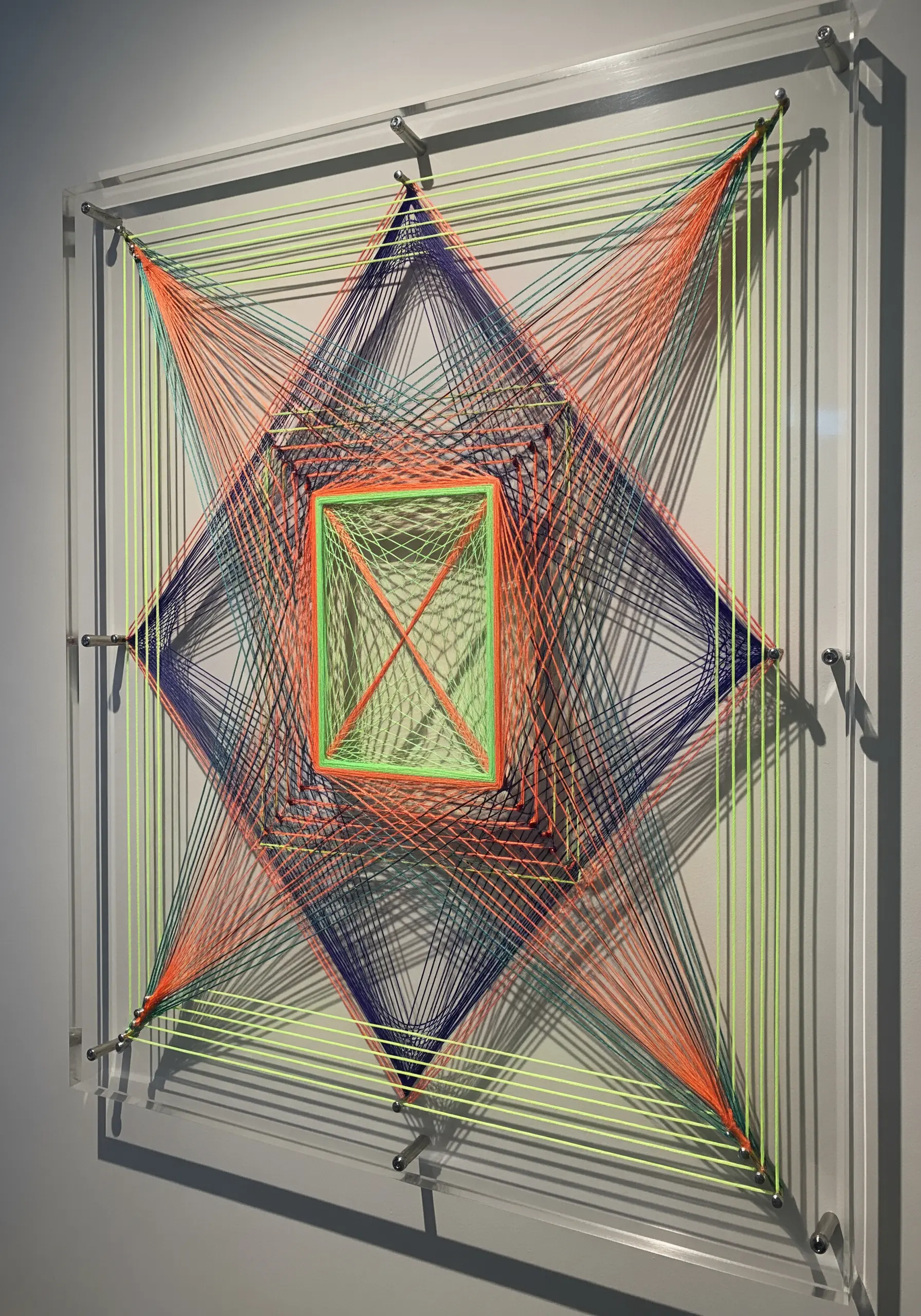 Geometric string art on a clear acrylic panel with layers of orange, blue, and neon green thread.
