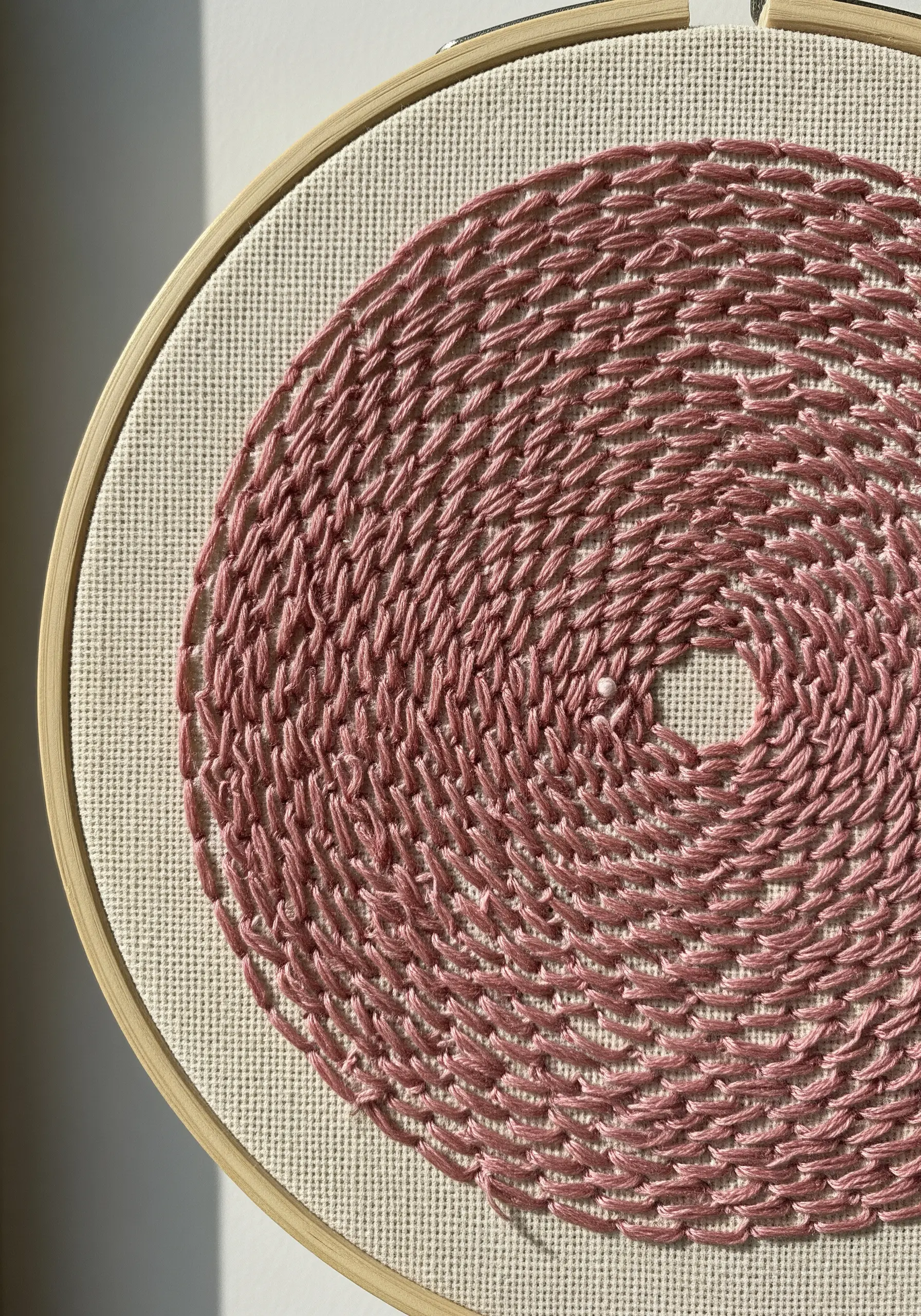 A circular spiral pattern filled with dense, randomly oriented seed stitches in dusty rose thread.