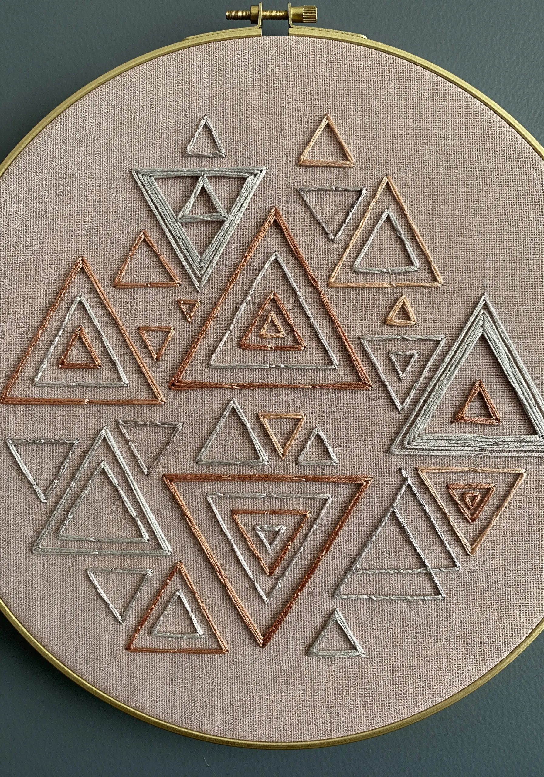 Nested geometric triangles stitched in silver and copper metallic thread on neutral fabric.