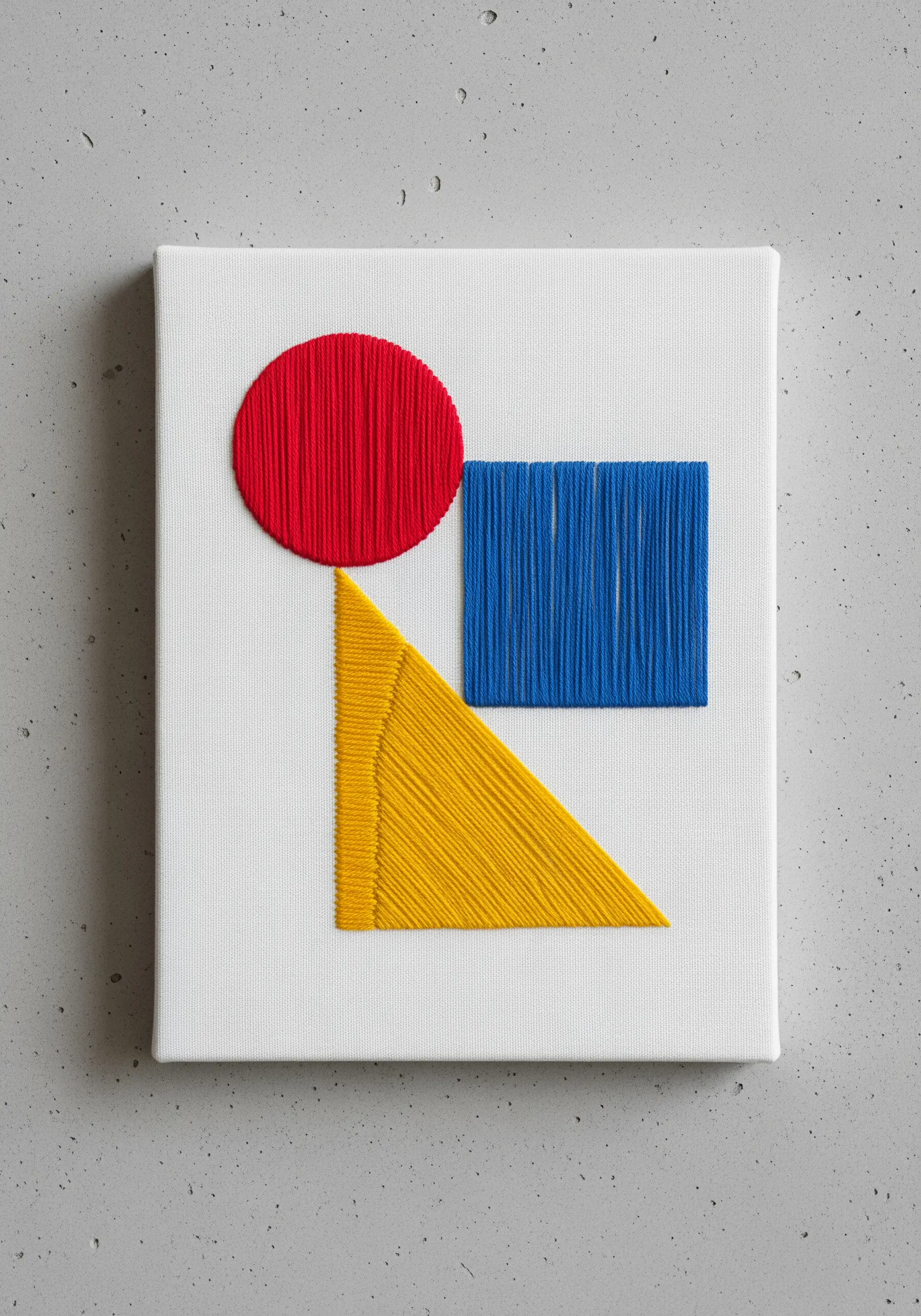 Geometric art with a red circle, blue square, and yellow triangle stitched on white canvas.