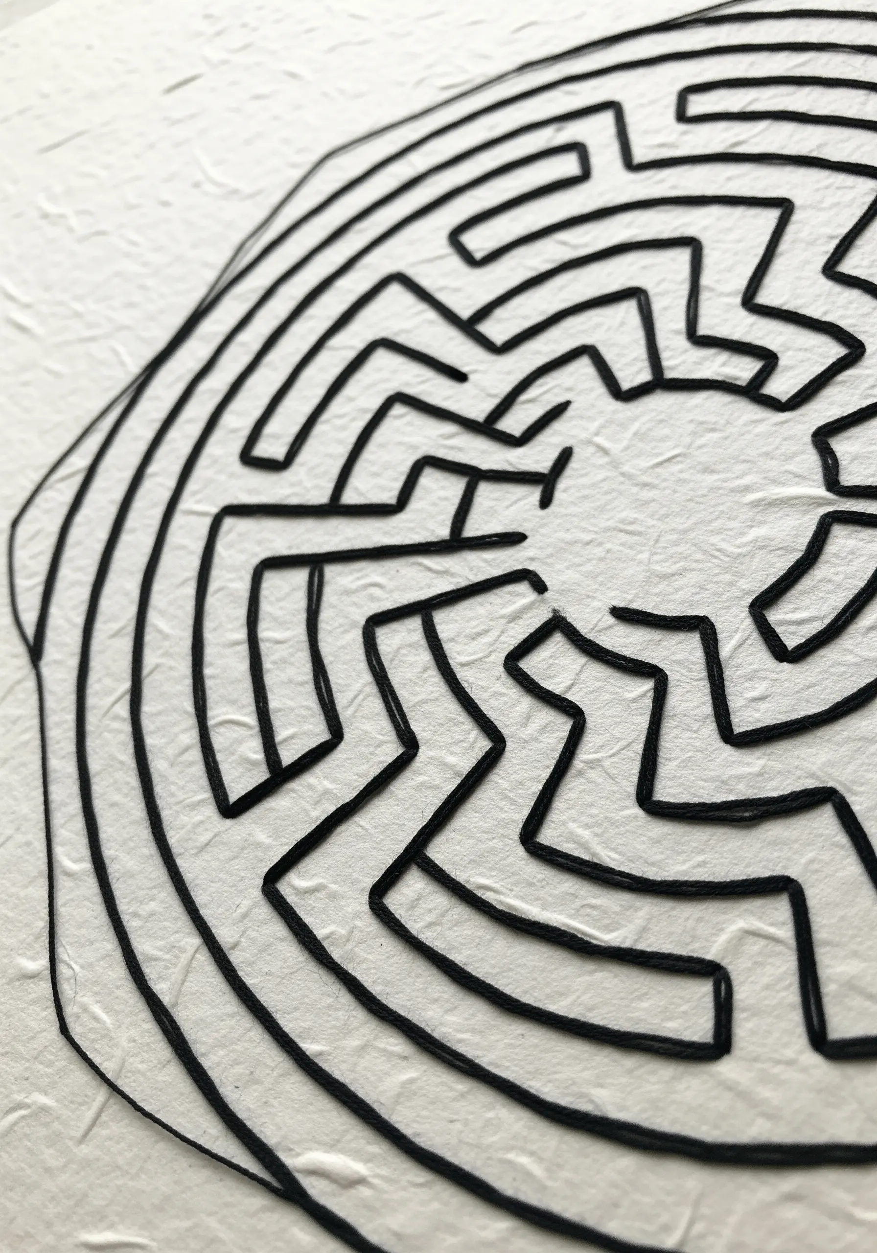 A black thread labyrinth design stitched onto textured, handmade white paper.