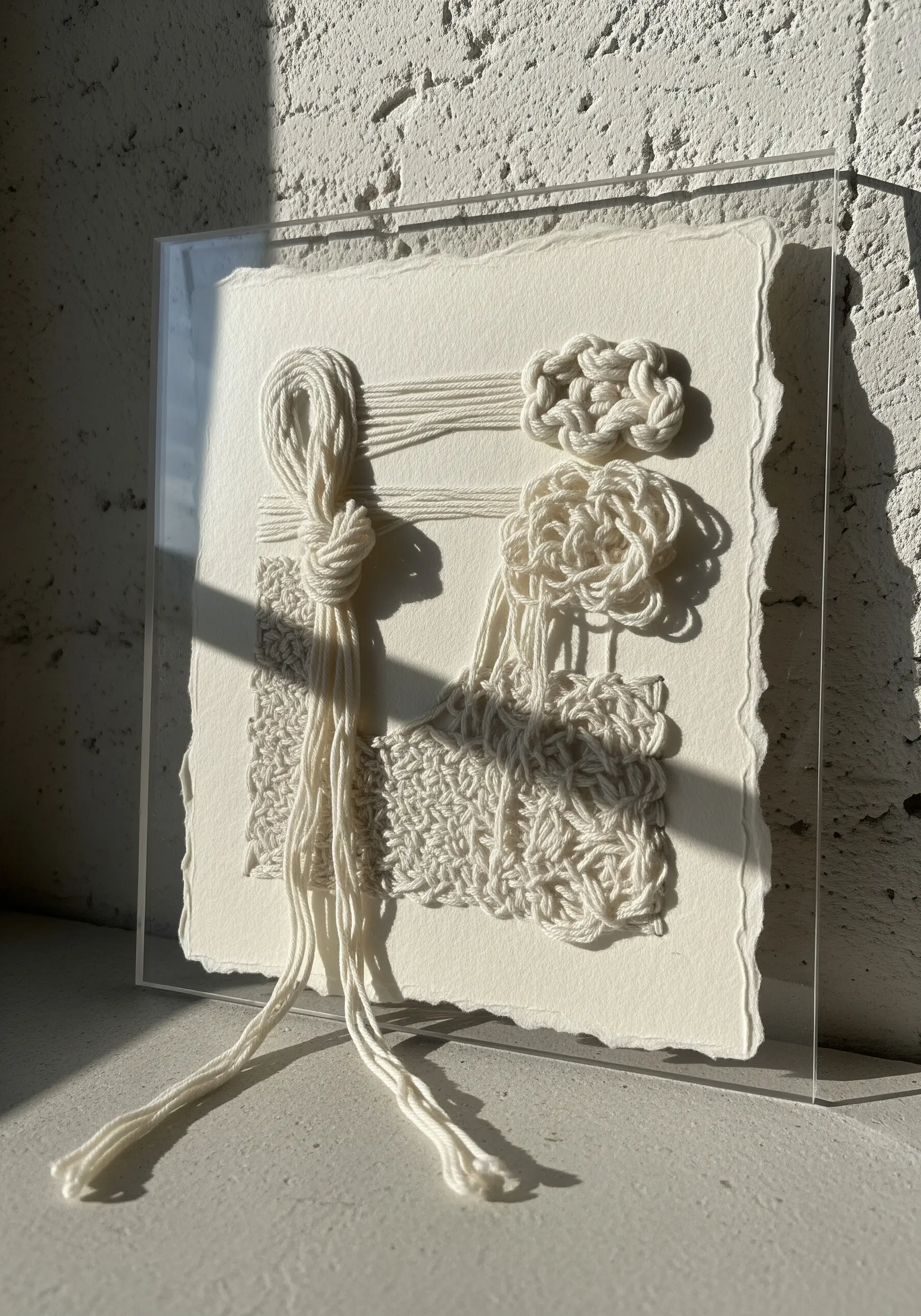 A monochrome white fiber art piece with knots and weaving on handmade paper.