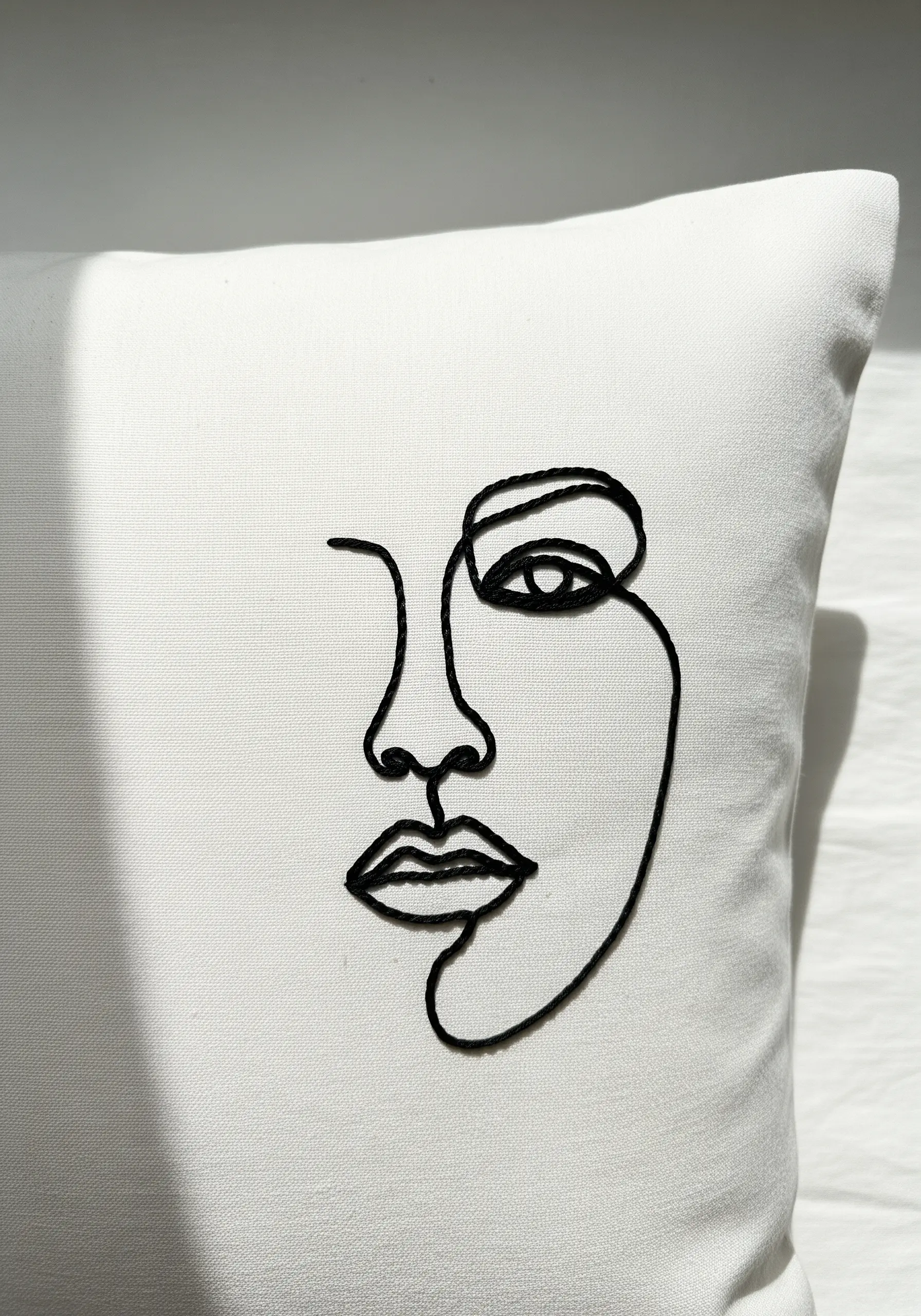 An abstract face rendered in a single, continuous black embroidered line on a white pillow.