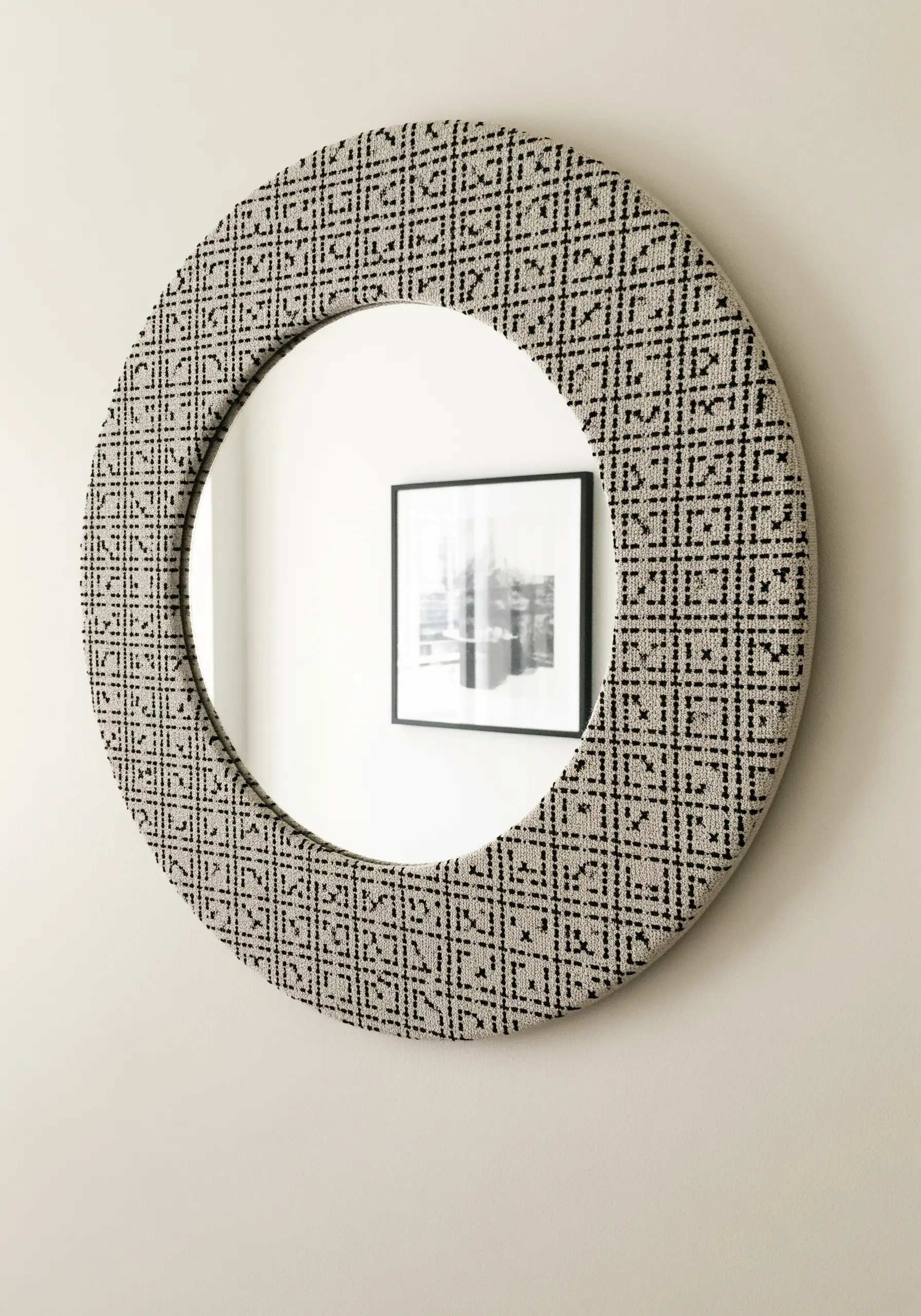 A round mirror with its frame covered in fabric embroidered with a black geometric pattern