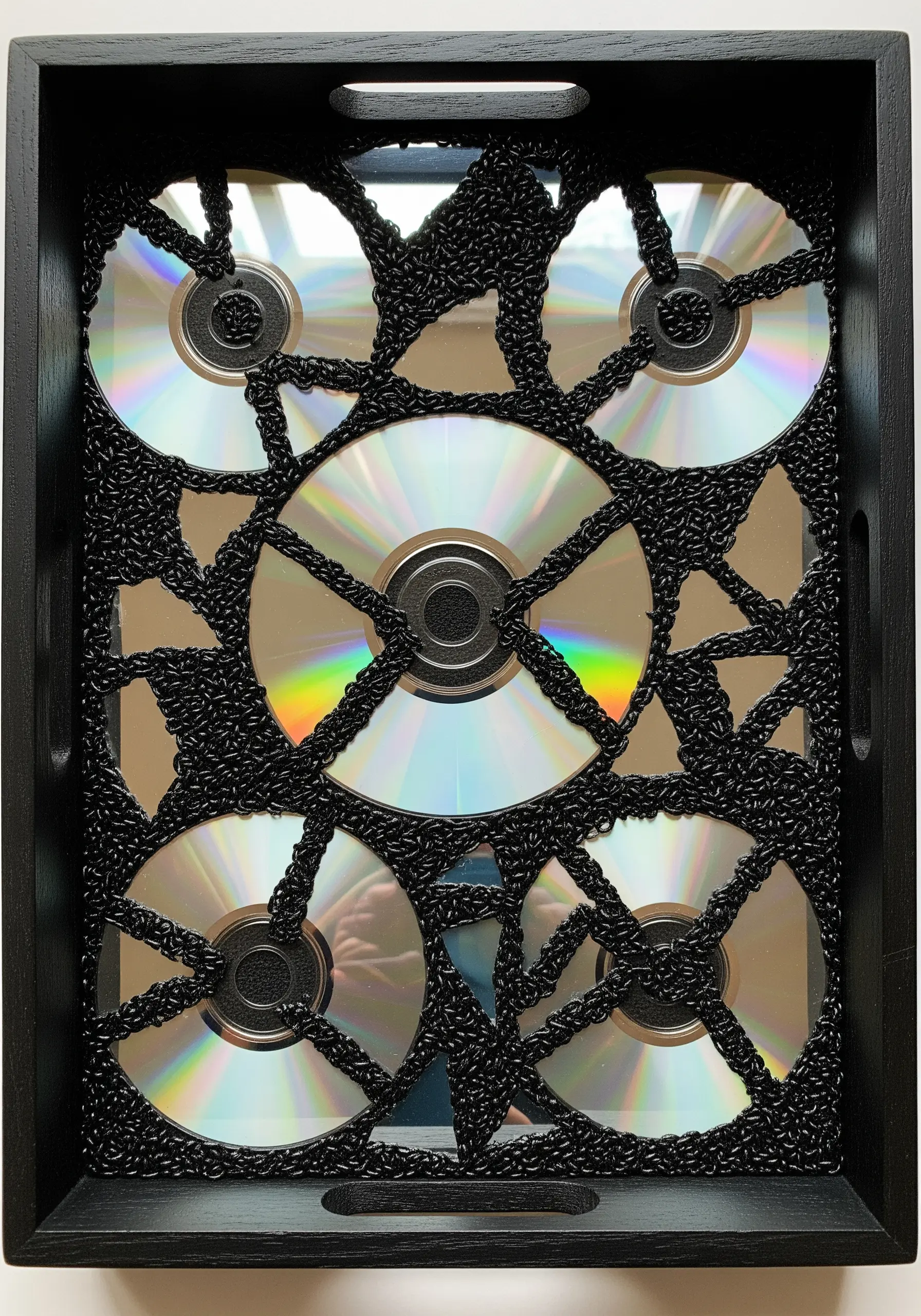 Old CDs arranged in a black tray, overlaid with a web of chunky black crocheted yarn.