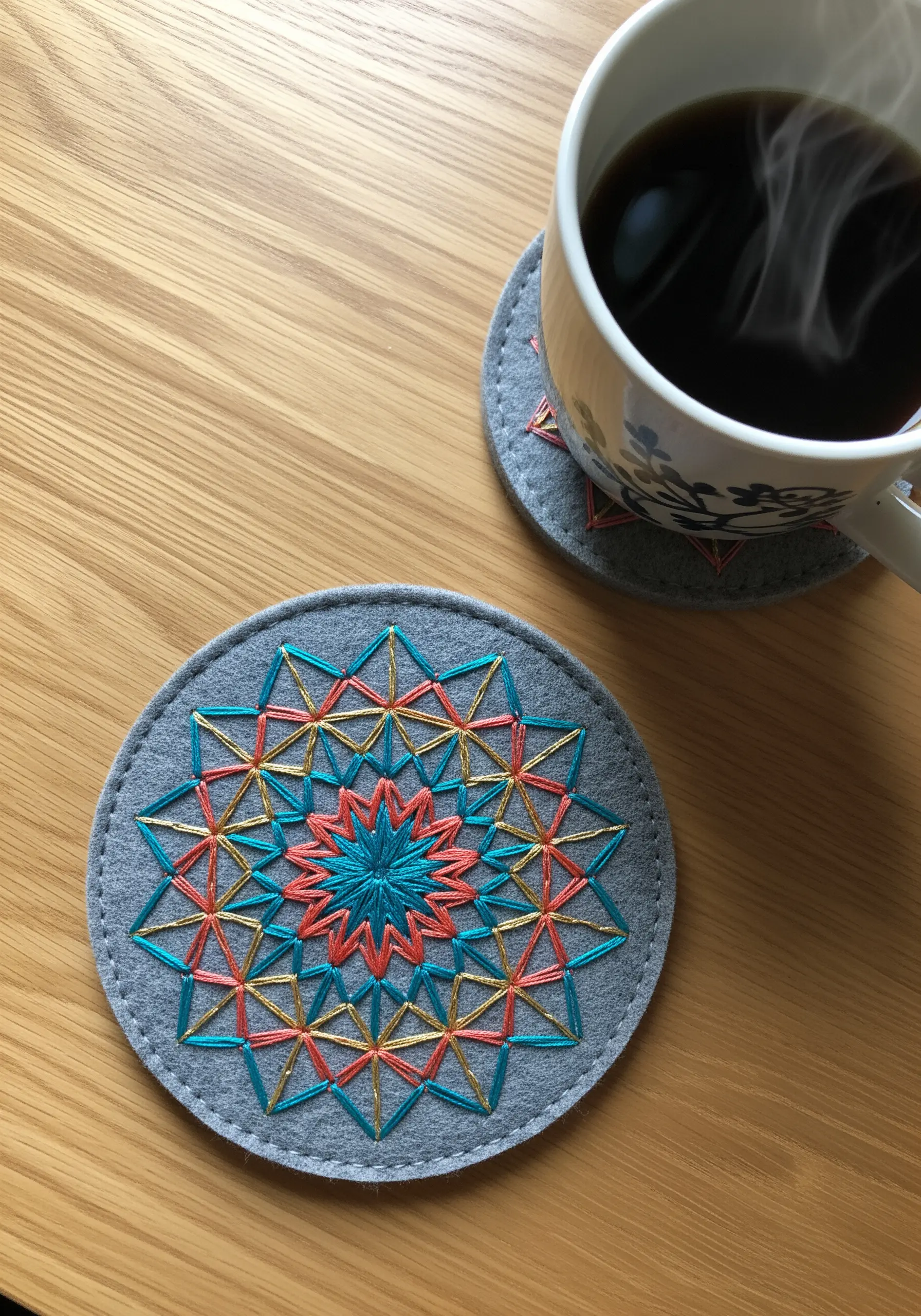Geometric starburst embroidery on a grey felt coaster with coral, teal, and gold thread.