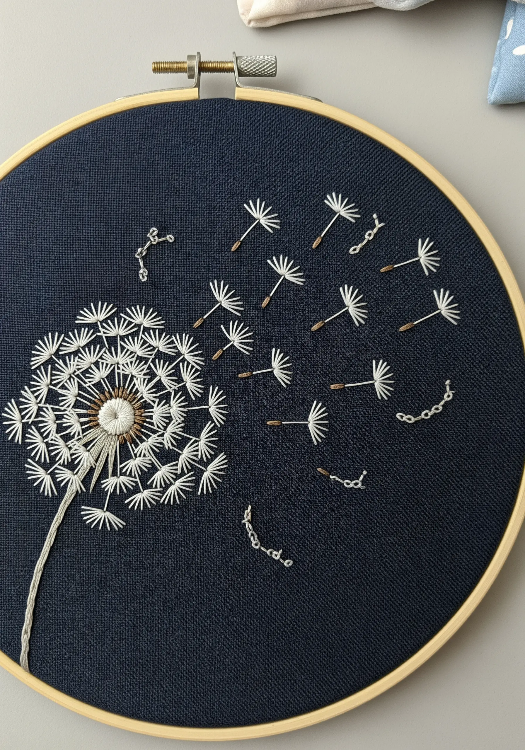A dandelion seed head embroidered on dark fabric, with seeds blowing away.