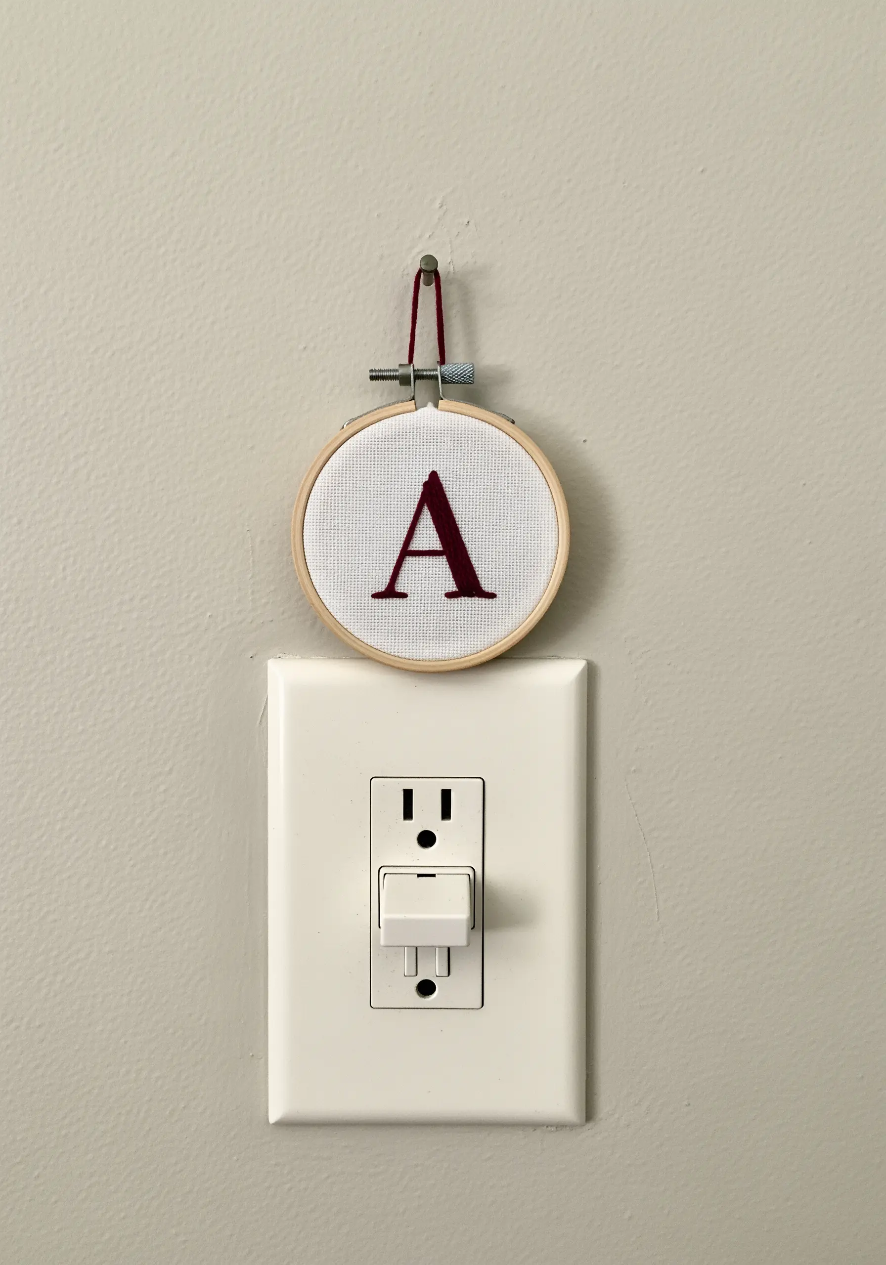 A bold, satin-stitched letter 'A' in a tiny hoop hanging above a light switch.