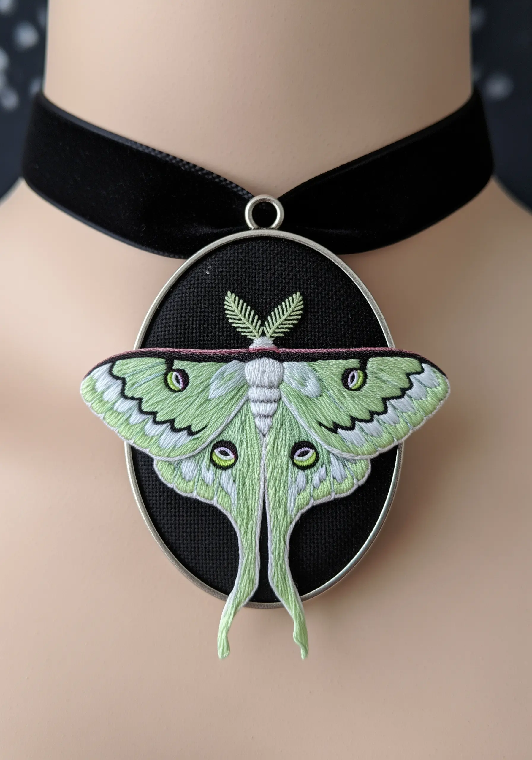An embroidered Luna moth pendant where the wings extend beyond the oval frame.