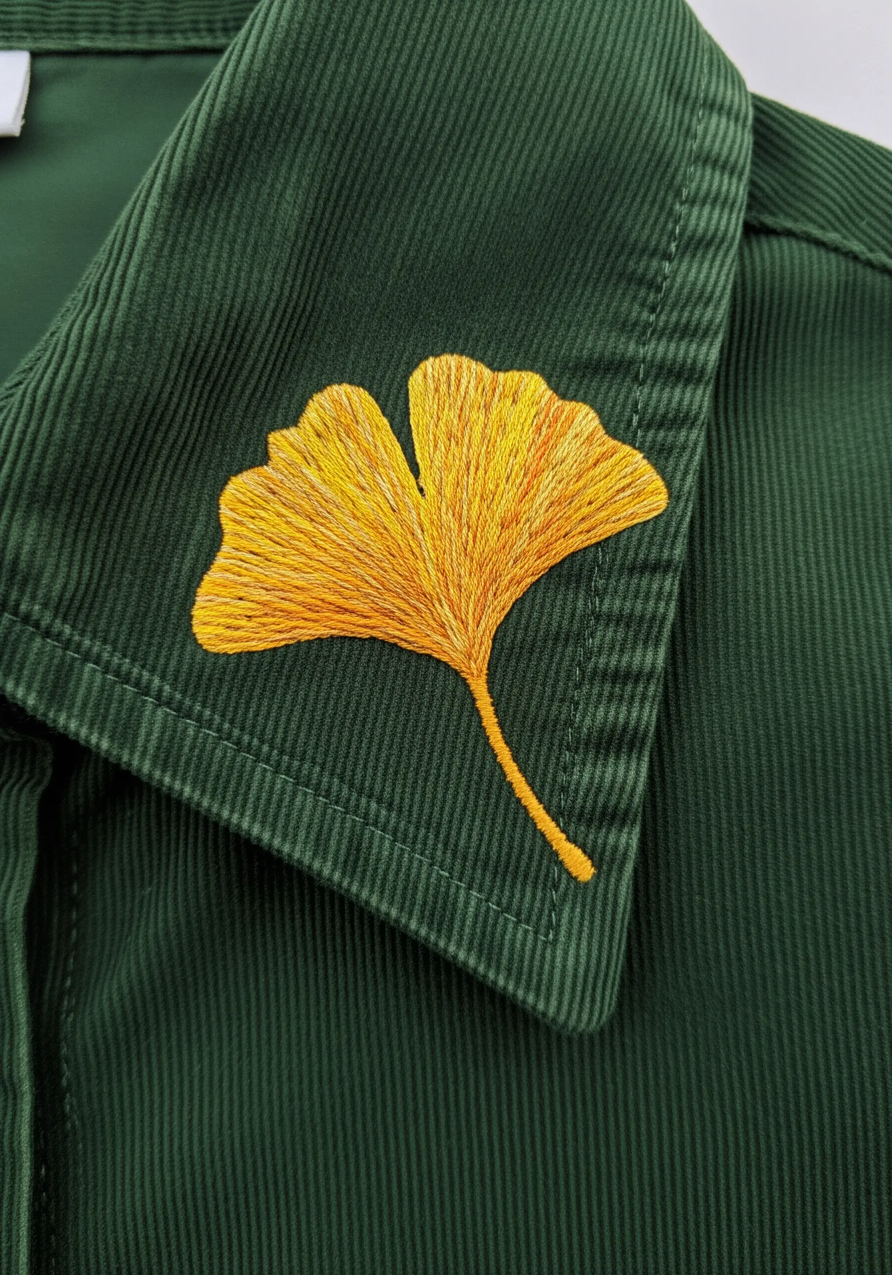A golden-yellow ginkgo leaf embroidered with long stitches on a dark green corduroy collar.