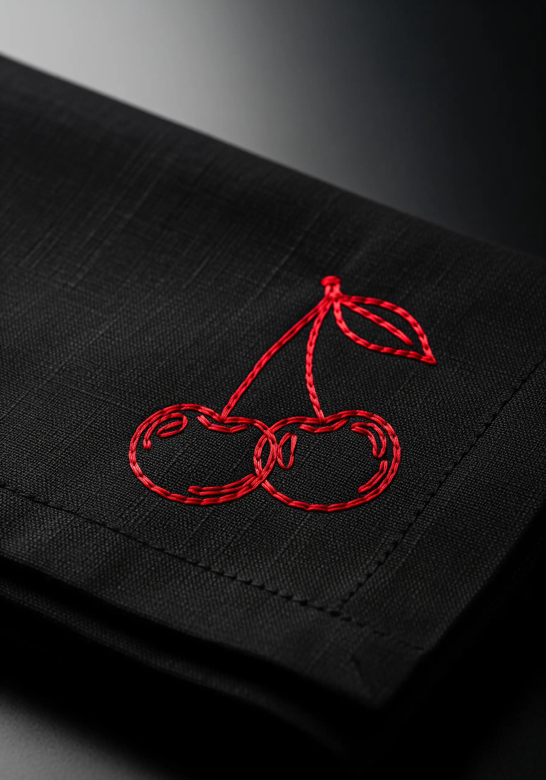 Two red cherries with intertwined stems embroidered on a black linen napkin.