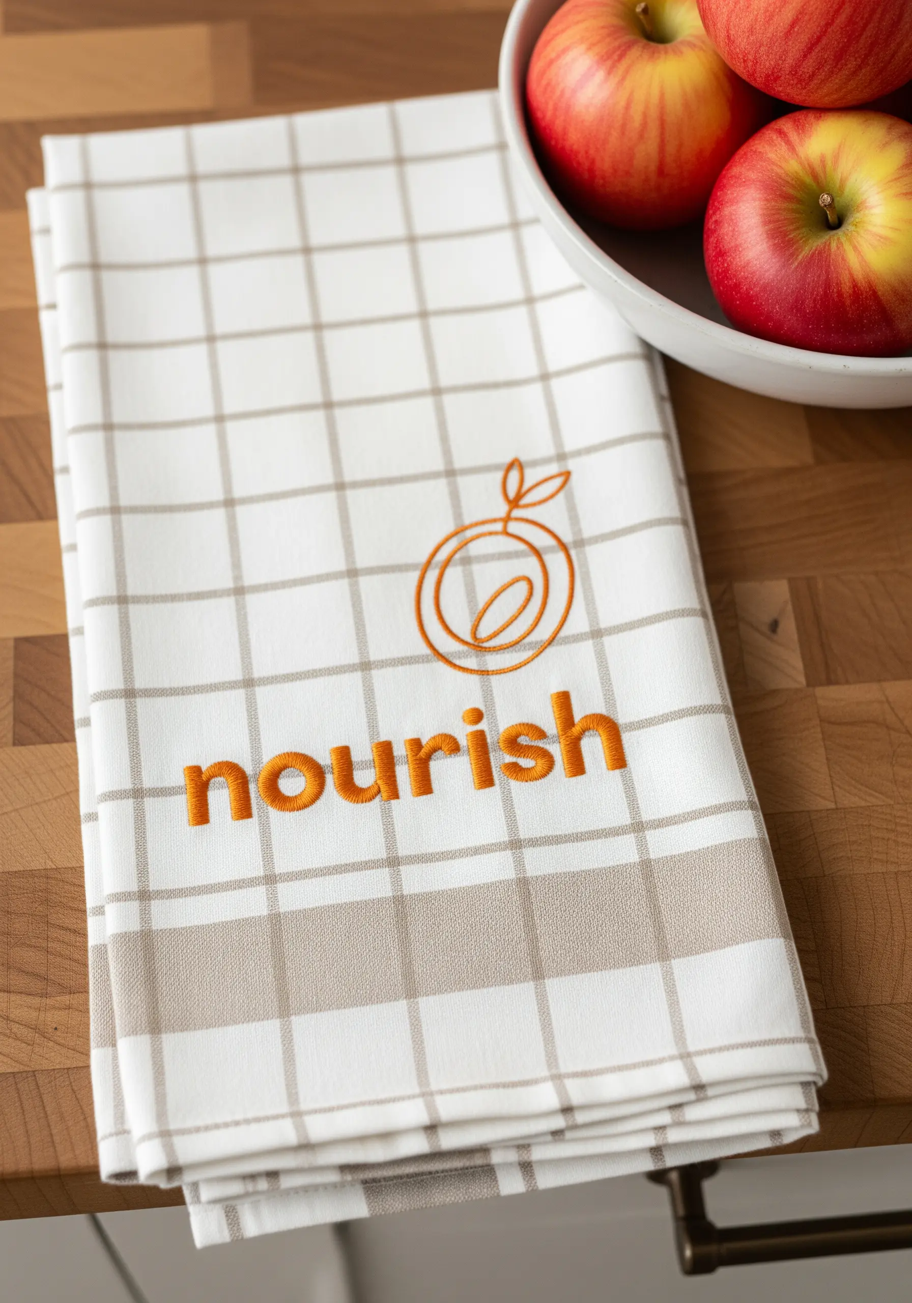 The word 'nourish' and a simple fruit icon embroidered in orange on a plaid kitchen towel