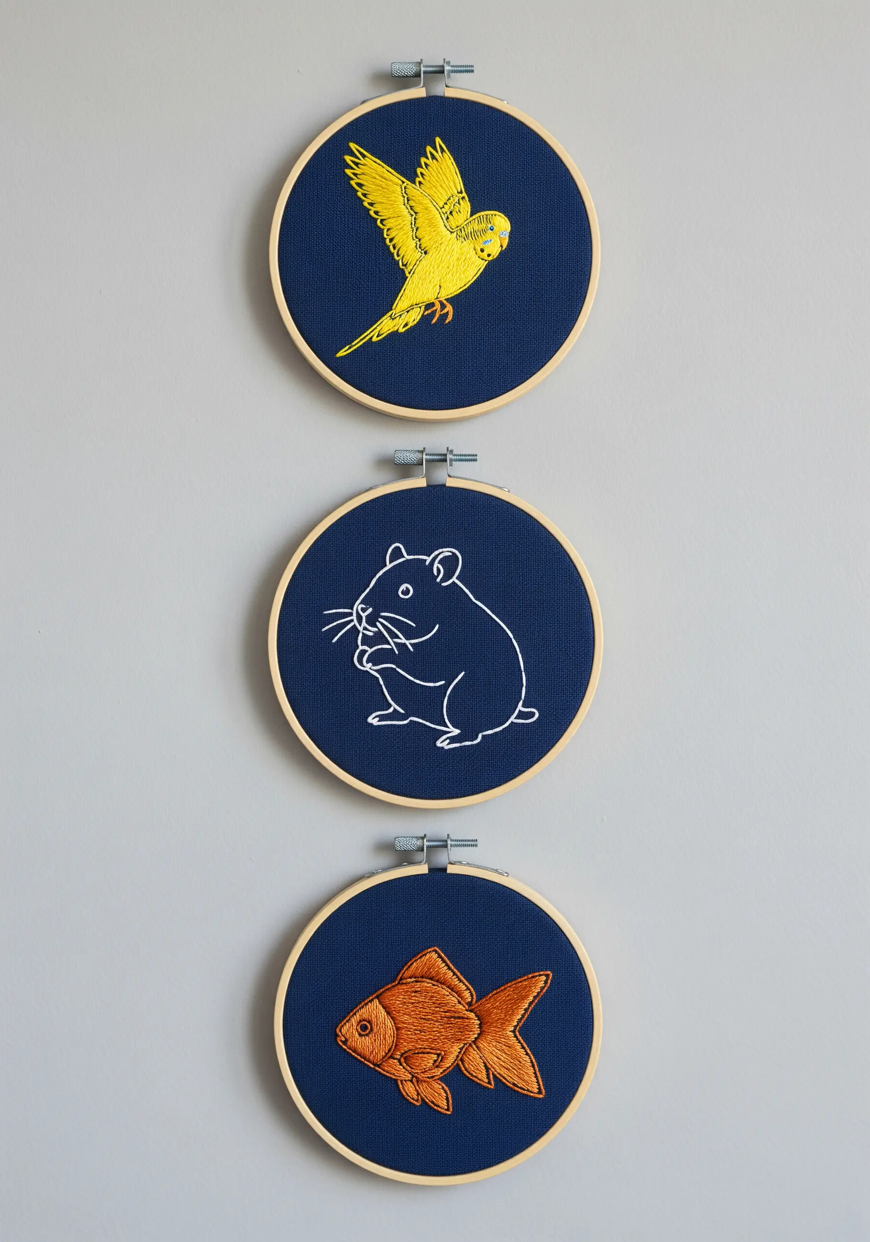Three embroidery hoops on a dark background: yellow bird, white hamster, orange fish.