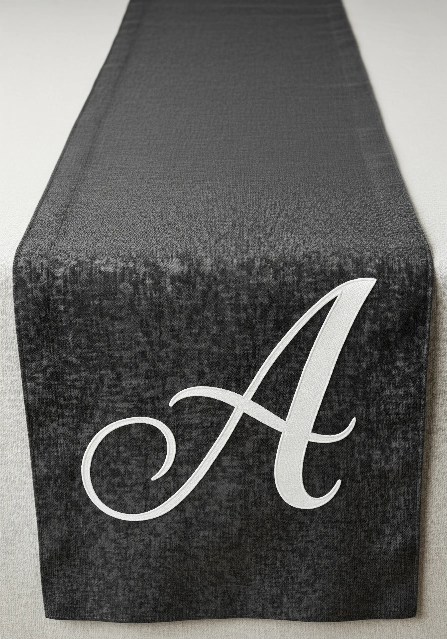 A large, elegant, white satin-stitched initial 'A' on a charcoal grey table runner.