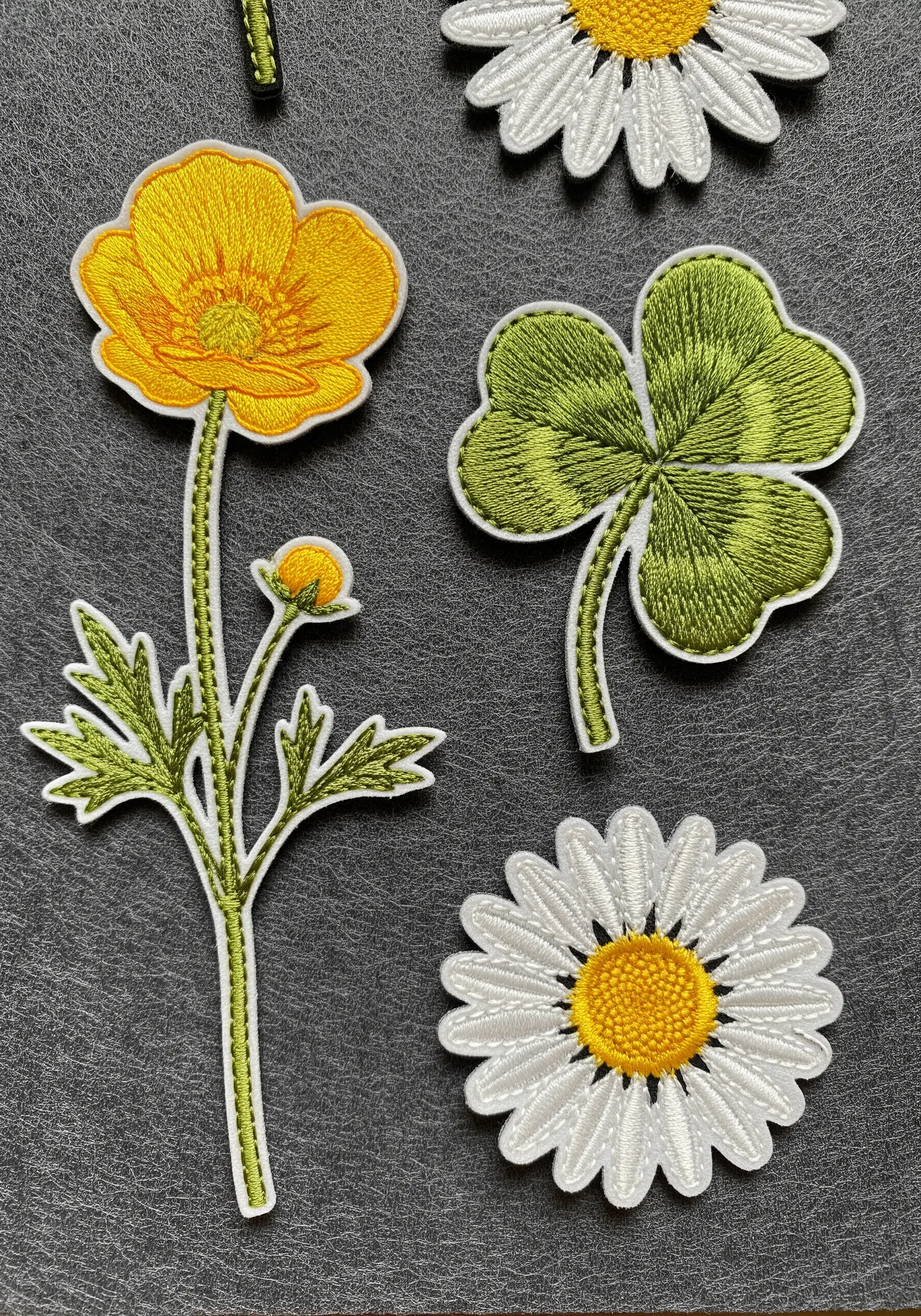 Hand-embroidered patches of a buttercup, clover, and daisy on a dark background.
