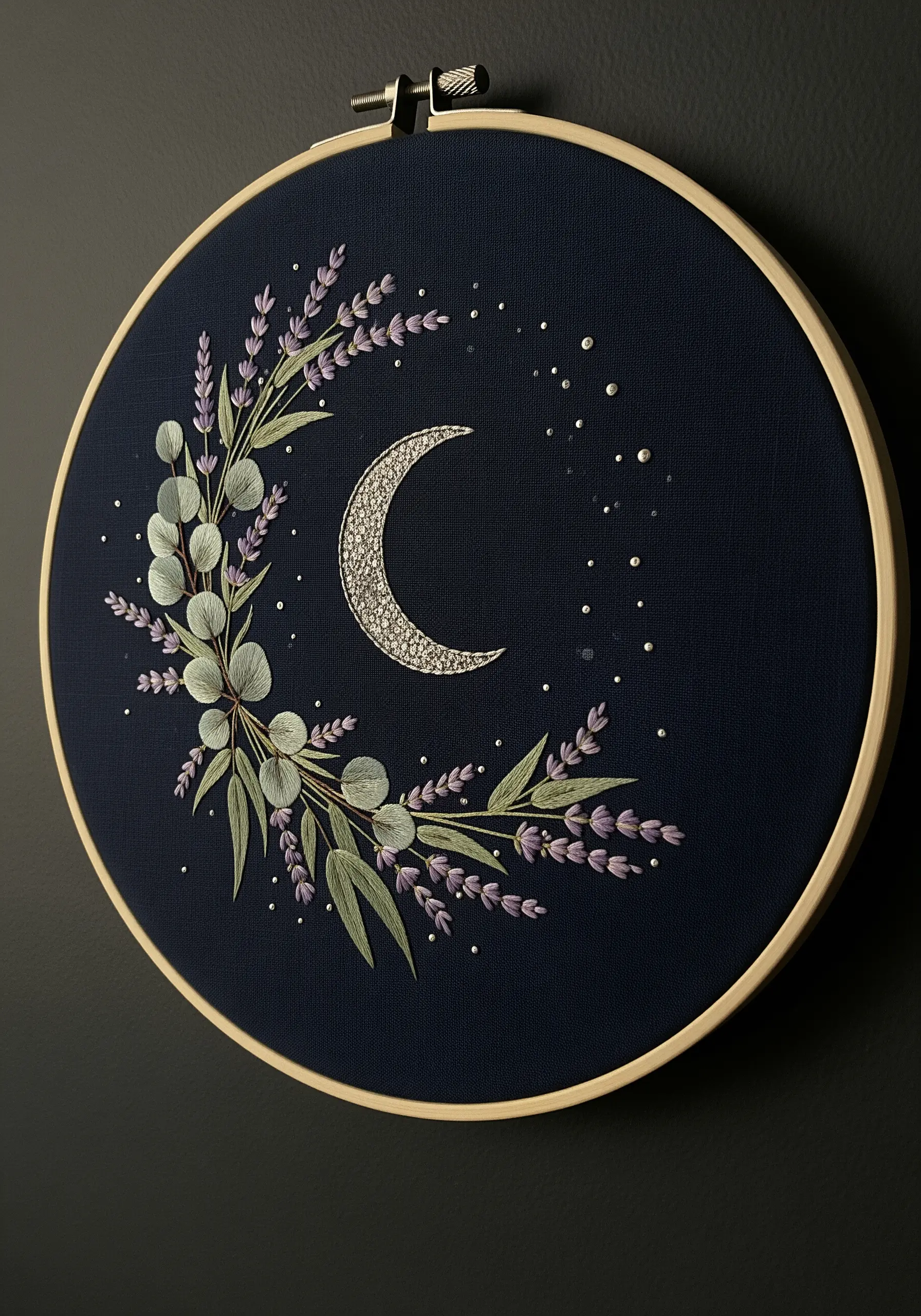 Embroidery hoop on dark fabric showing a silver crescent moon with lavender and eucalyptus.