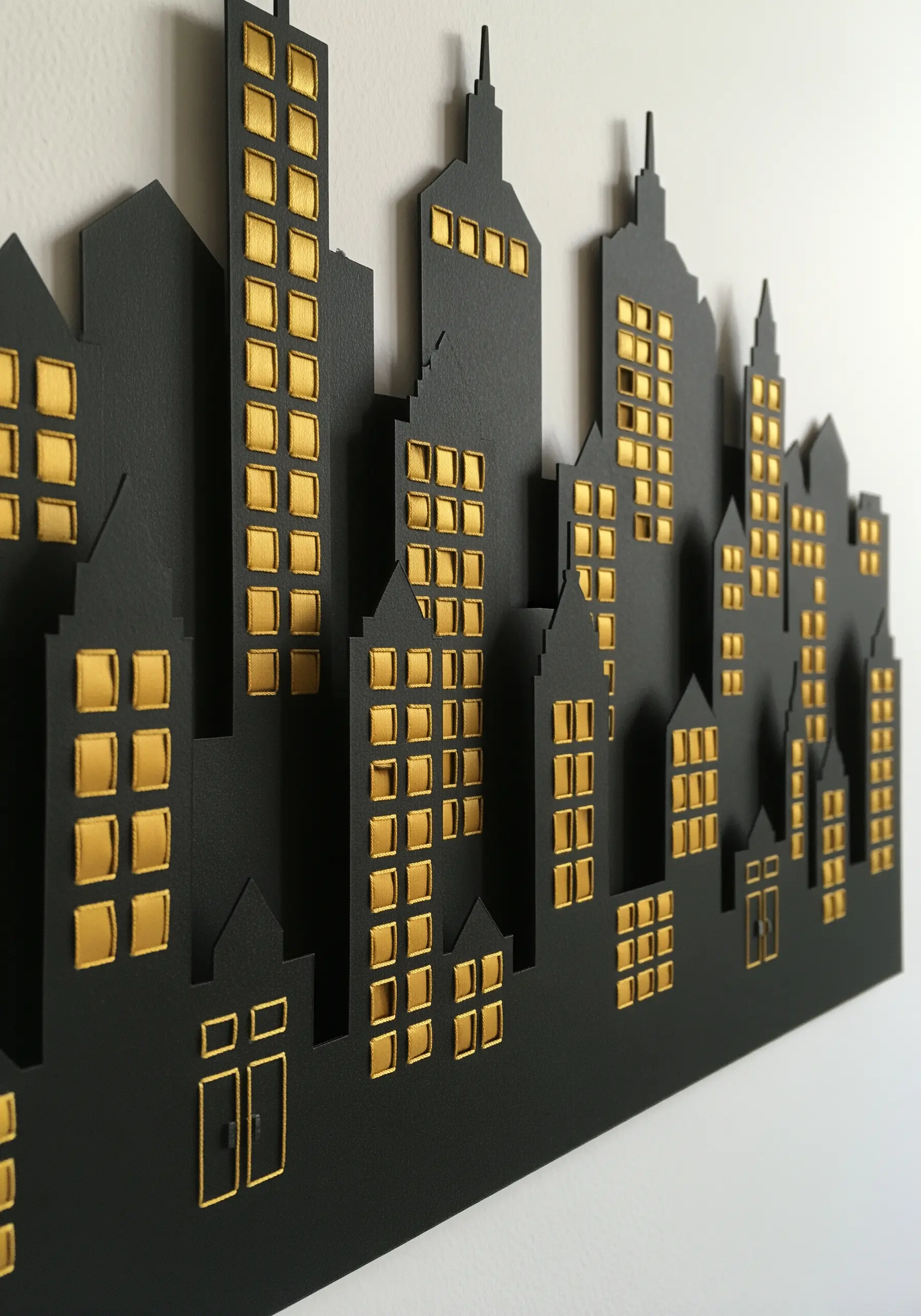 A black paper cityscape silhouette with windows filled in with shimmering gold satin stitch embroidery.
