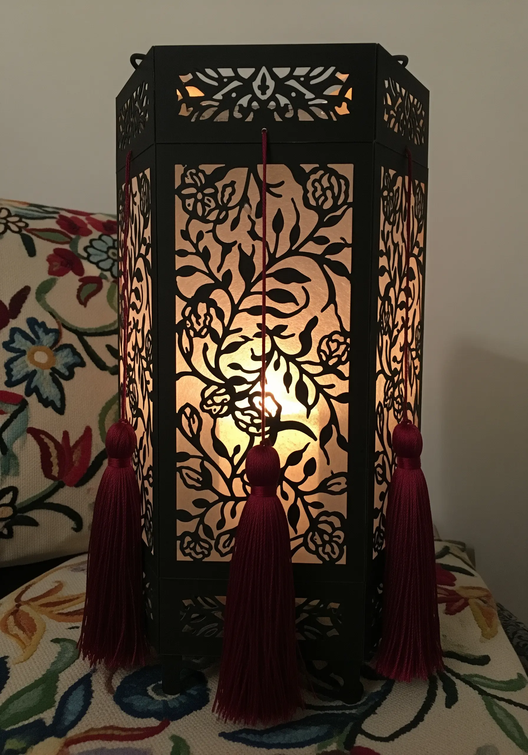 A black paper-cut lantern with floral motifs, lit from within and adorned with red tassels.