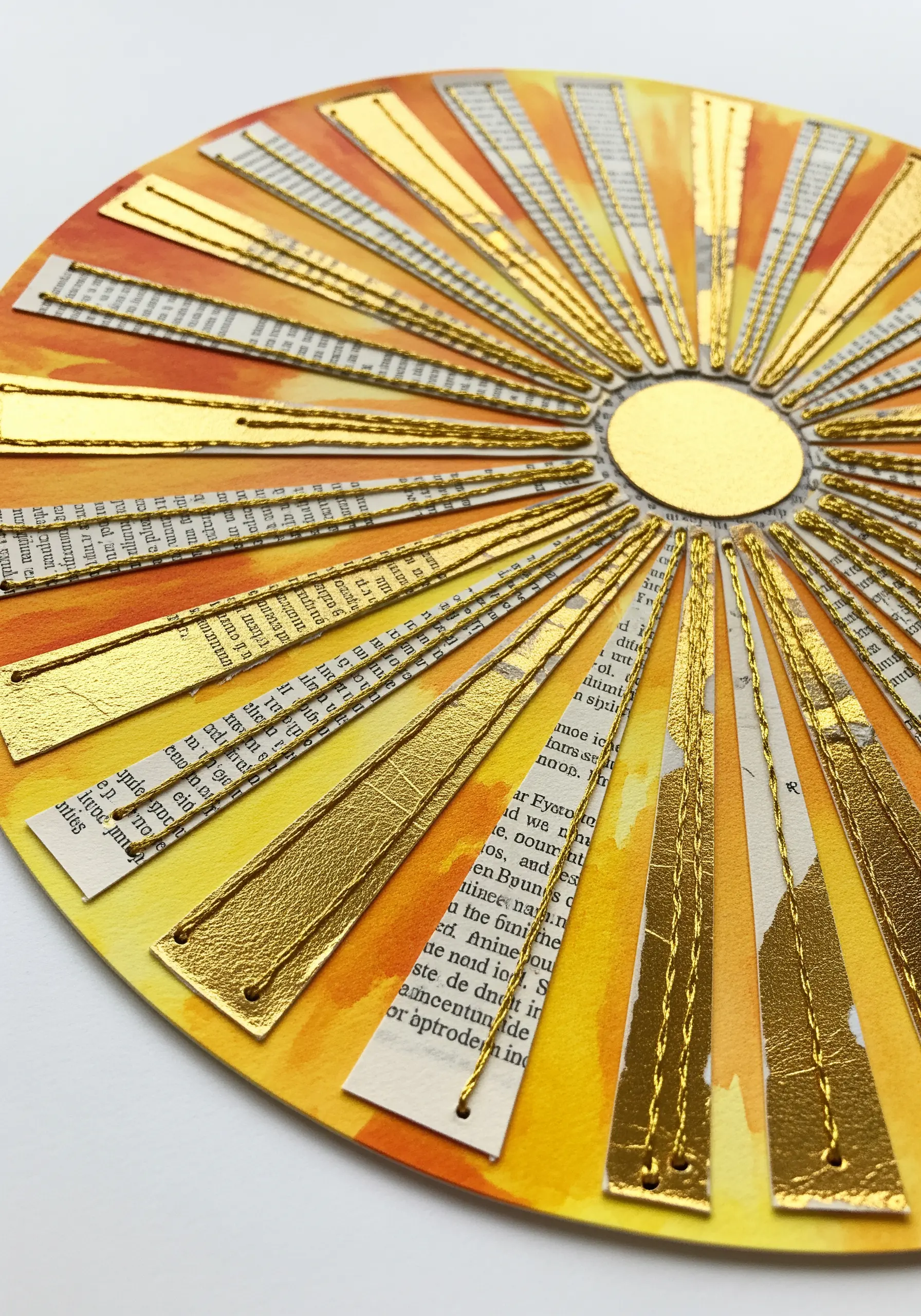 A sunburst collage made from watercolor paper, book pages, and metallic strips stitched with gold thread.