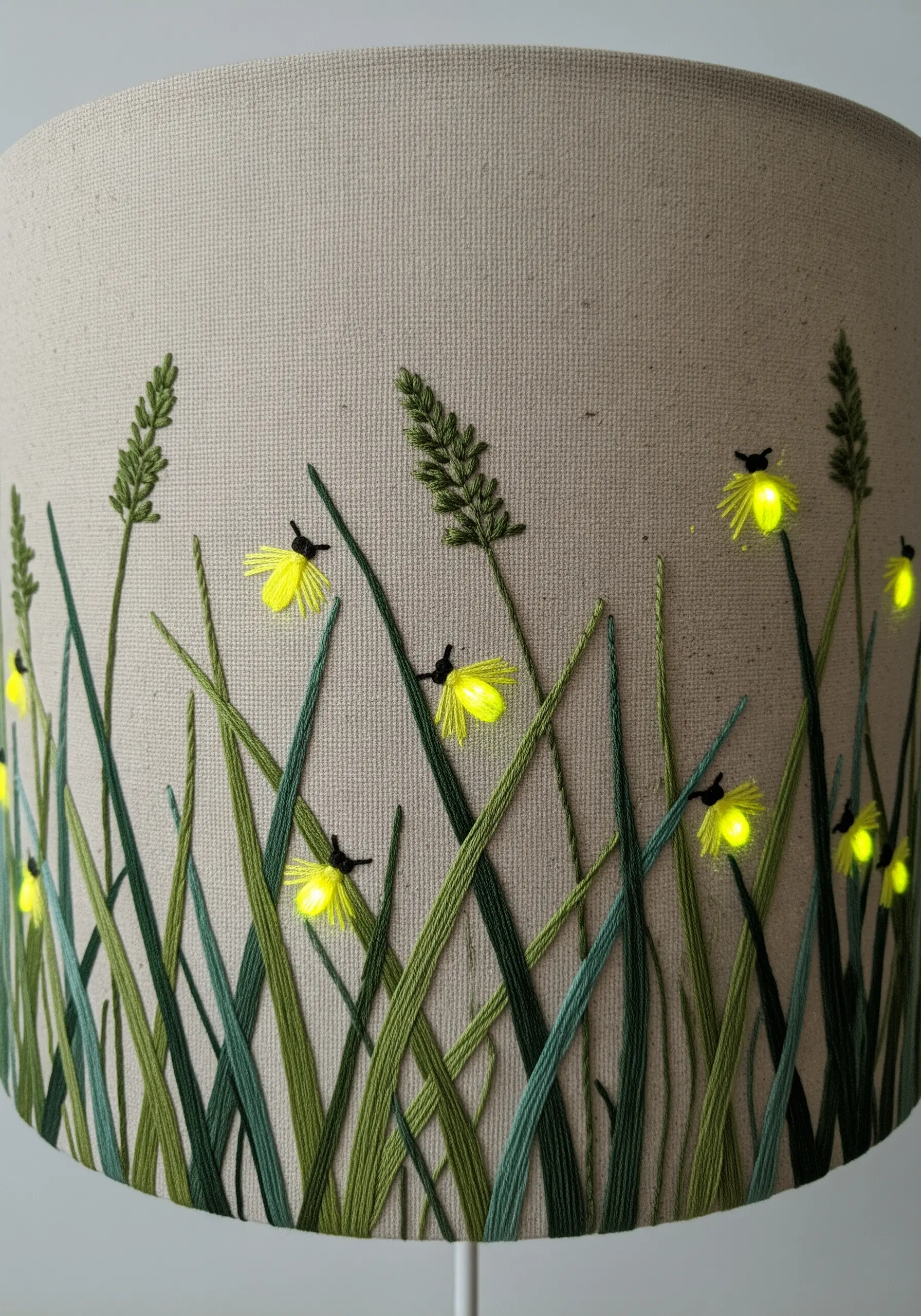 A lampshade embroidered with blades of grass and glowing fireflies lit by tiny LED lights.