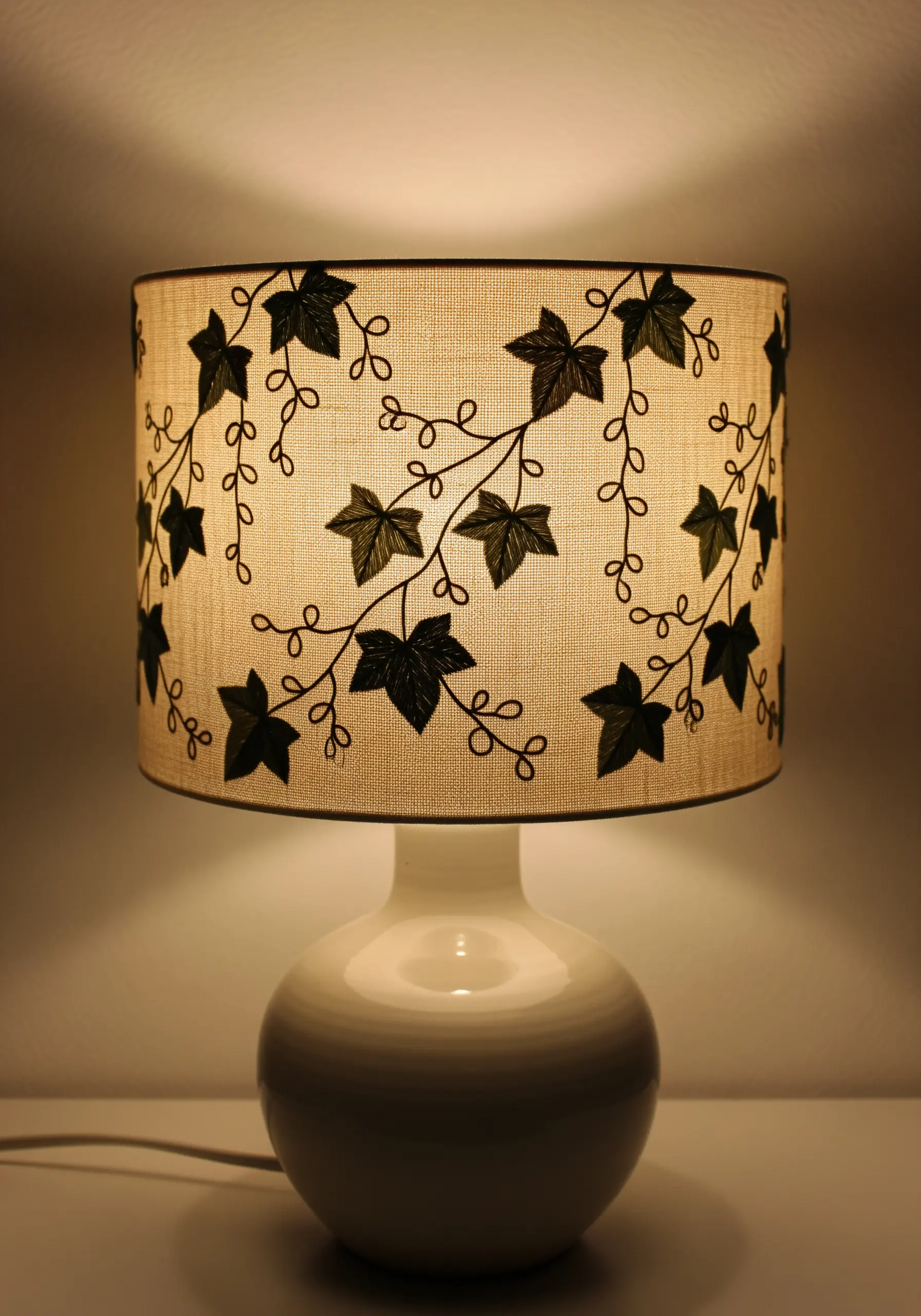 An illuminated drum lampshade with a trailing black ivy vine embroidered on it.