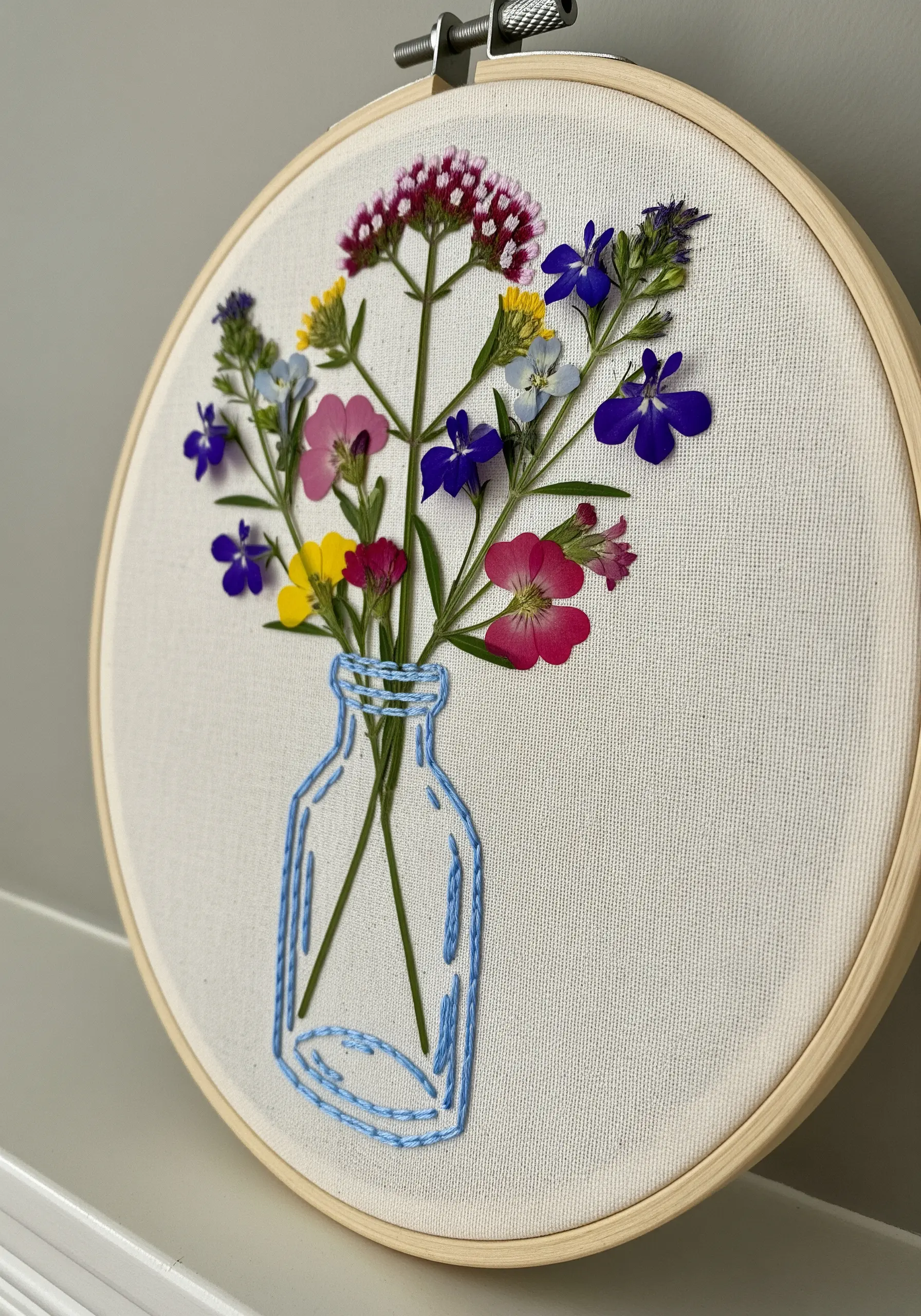 A line-art embroidery of a glass jar holding a bouquet of real pressed wildflowers.