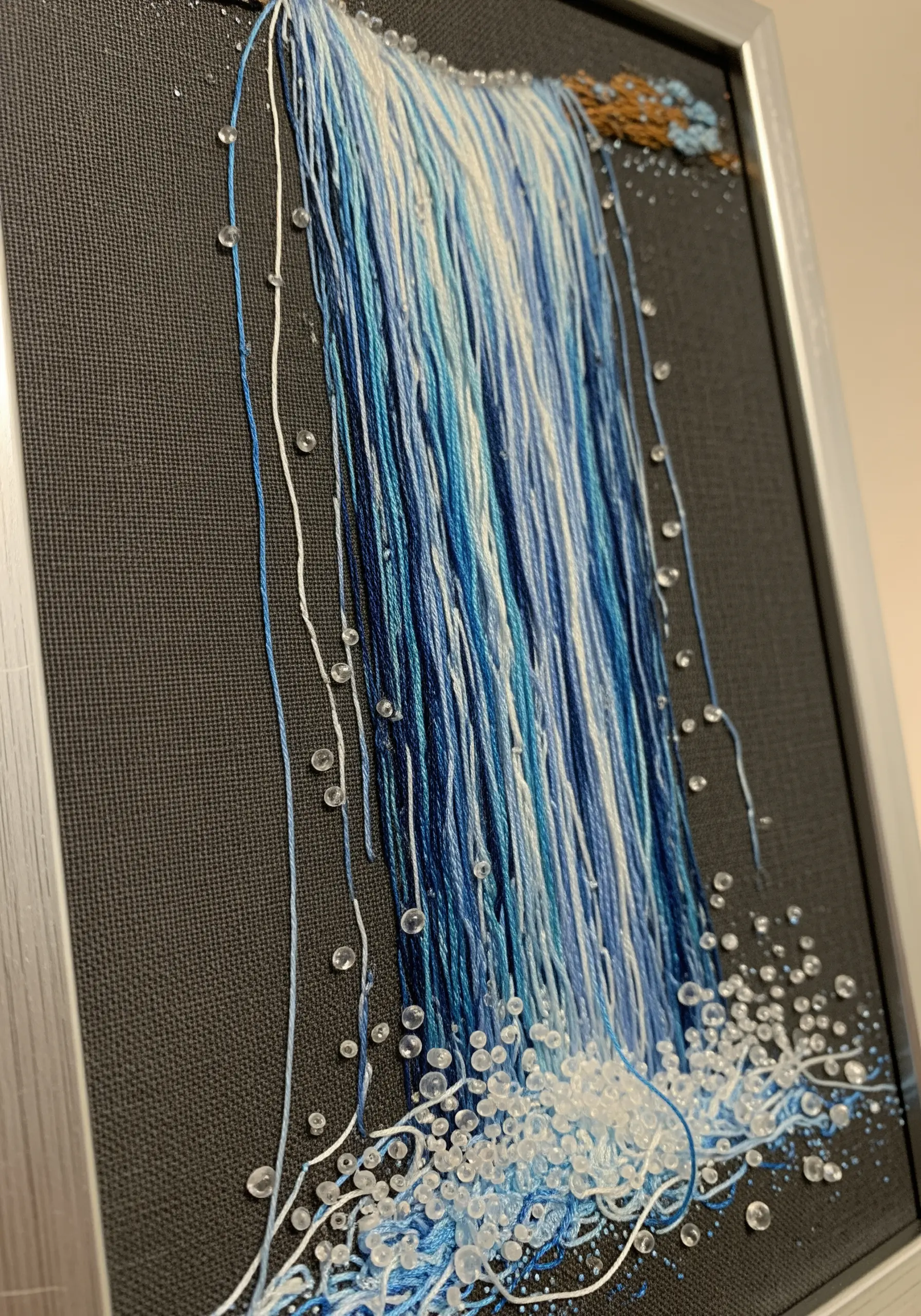 Embroidery of a waterfall using threads left loose to cascade down the fabric, with beads for spray.