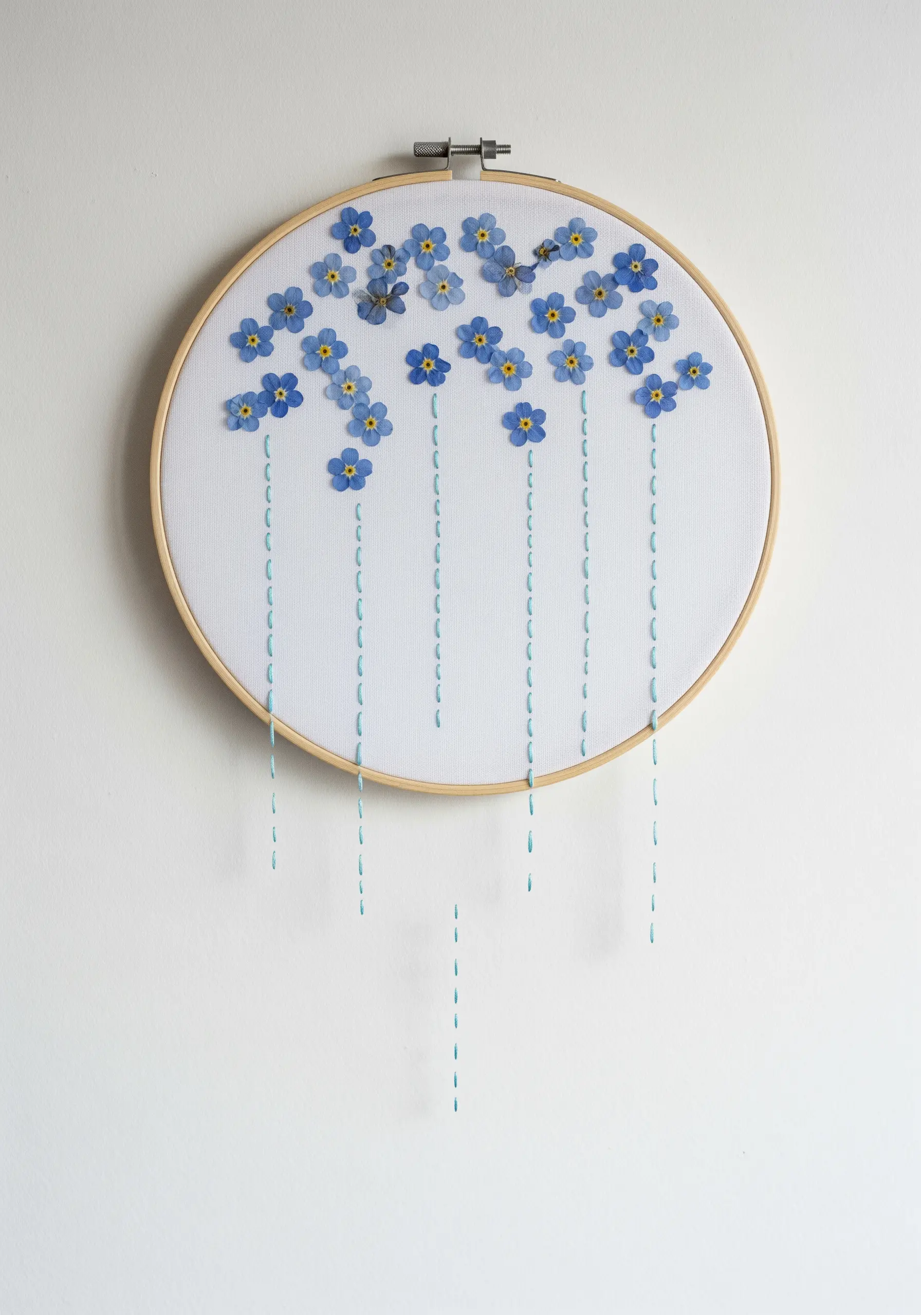Pressed forget-me-not flowers at the top of a hoop with simple, vertical stitch lines dripping down.