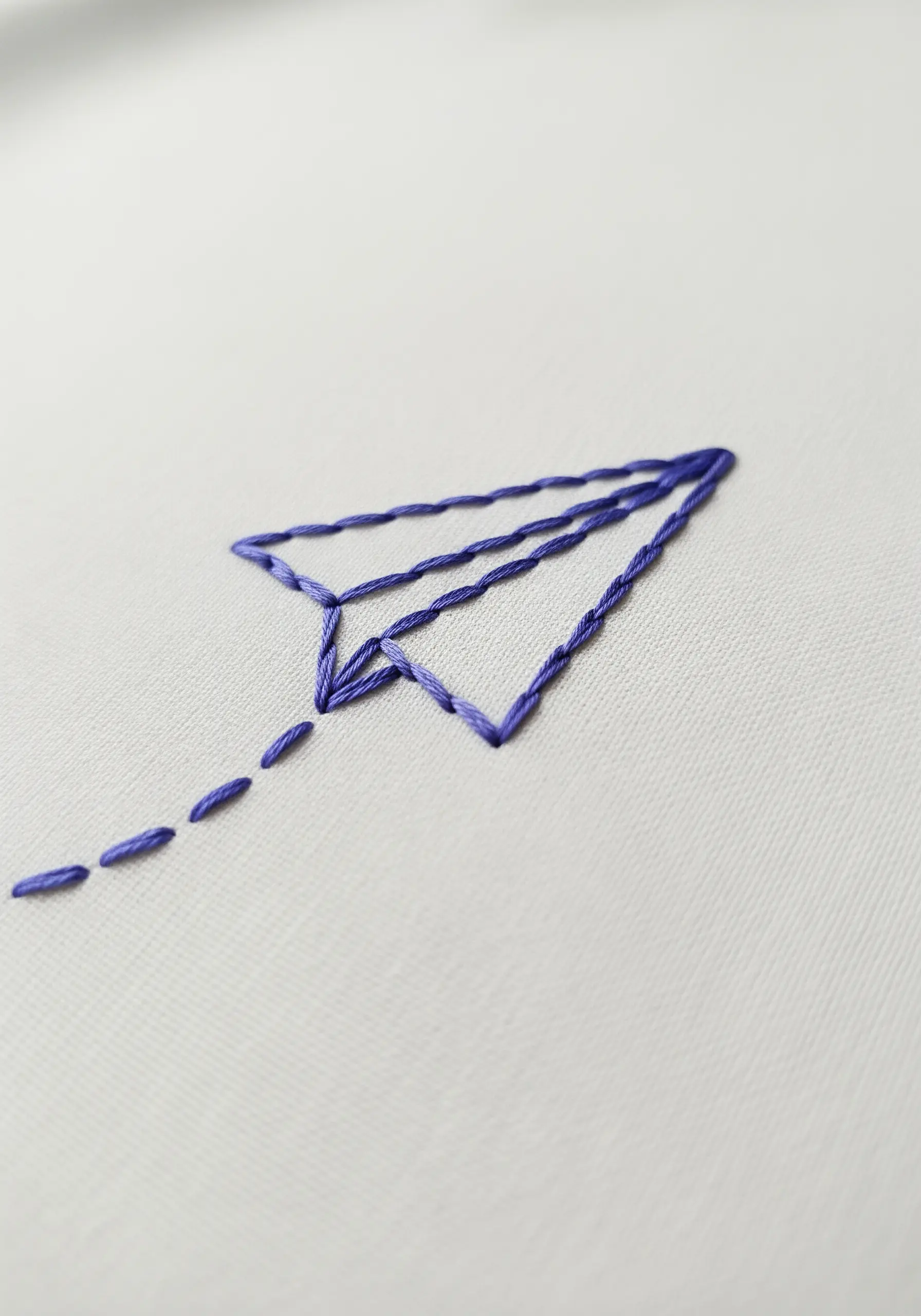 A simple outline of a paper airplane in purple thread with a dashed trail behind it.