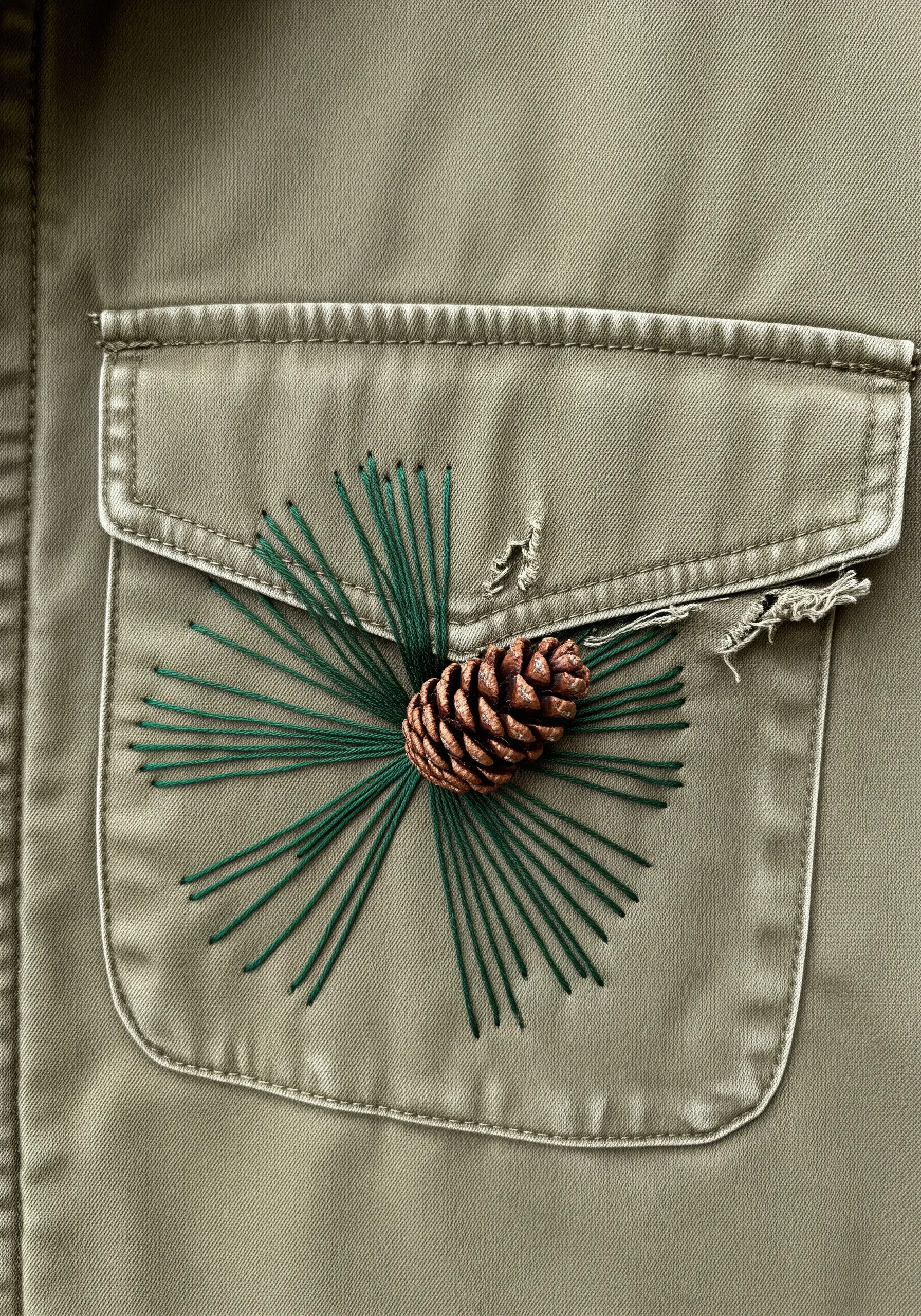 A real miniature pinecone stitched onto a shirt pocket with embroidered green pine needles radiating outward.