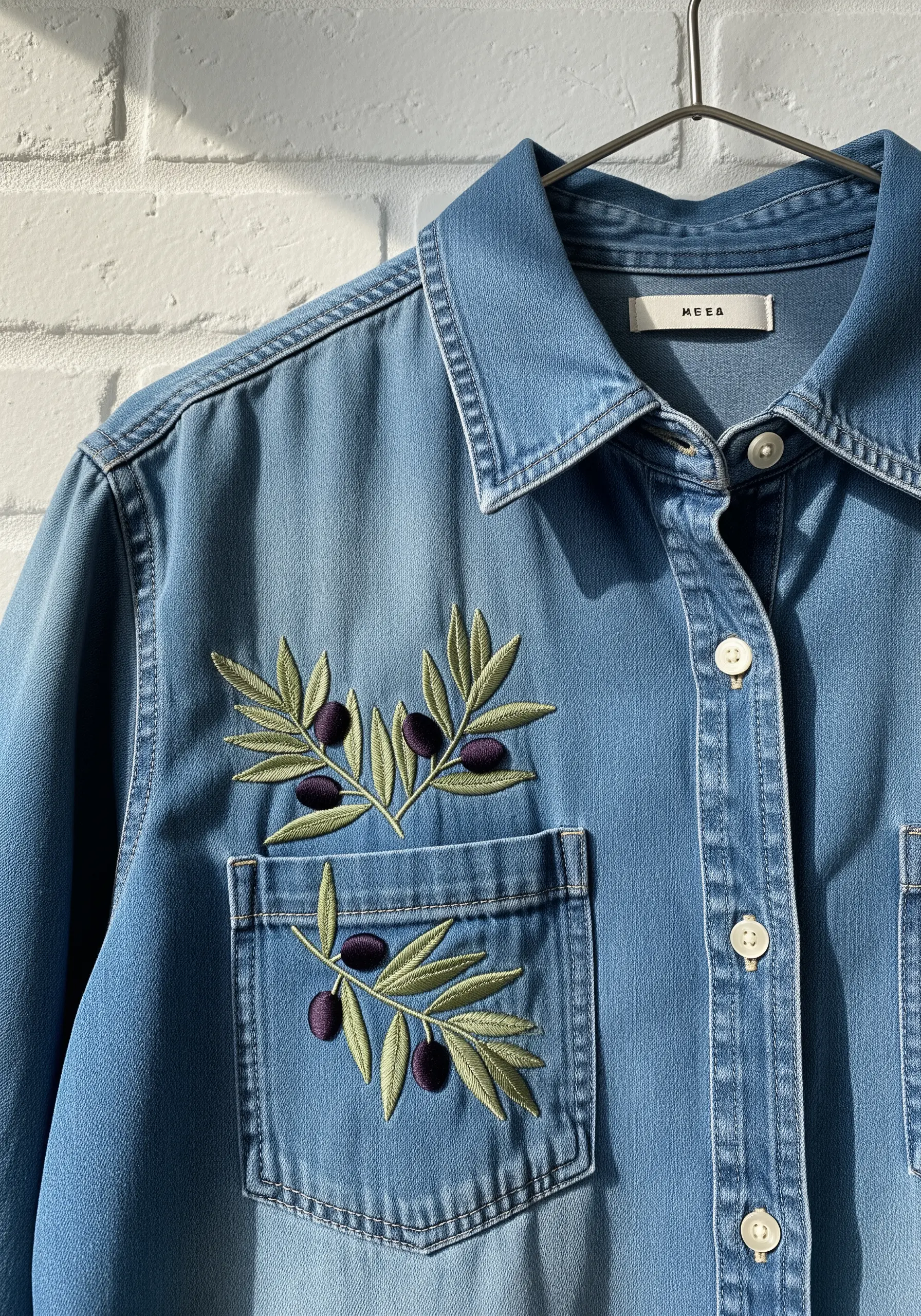 An olive branch with green leaves and purple olives stitched on and around a denim shirt pocket.