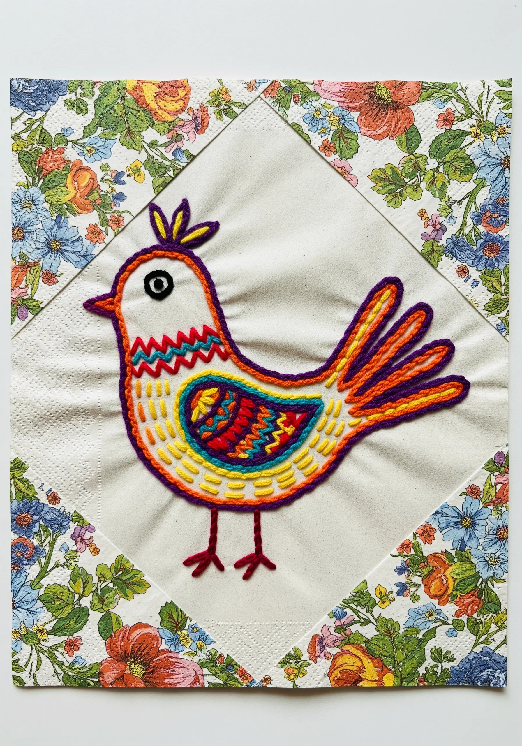 An embroidered bird on a paper napkin, decoupaged onto a canvas with a floral border.