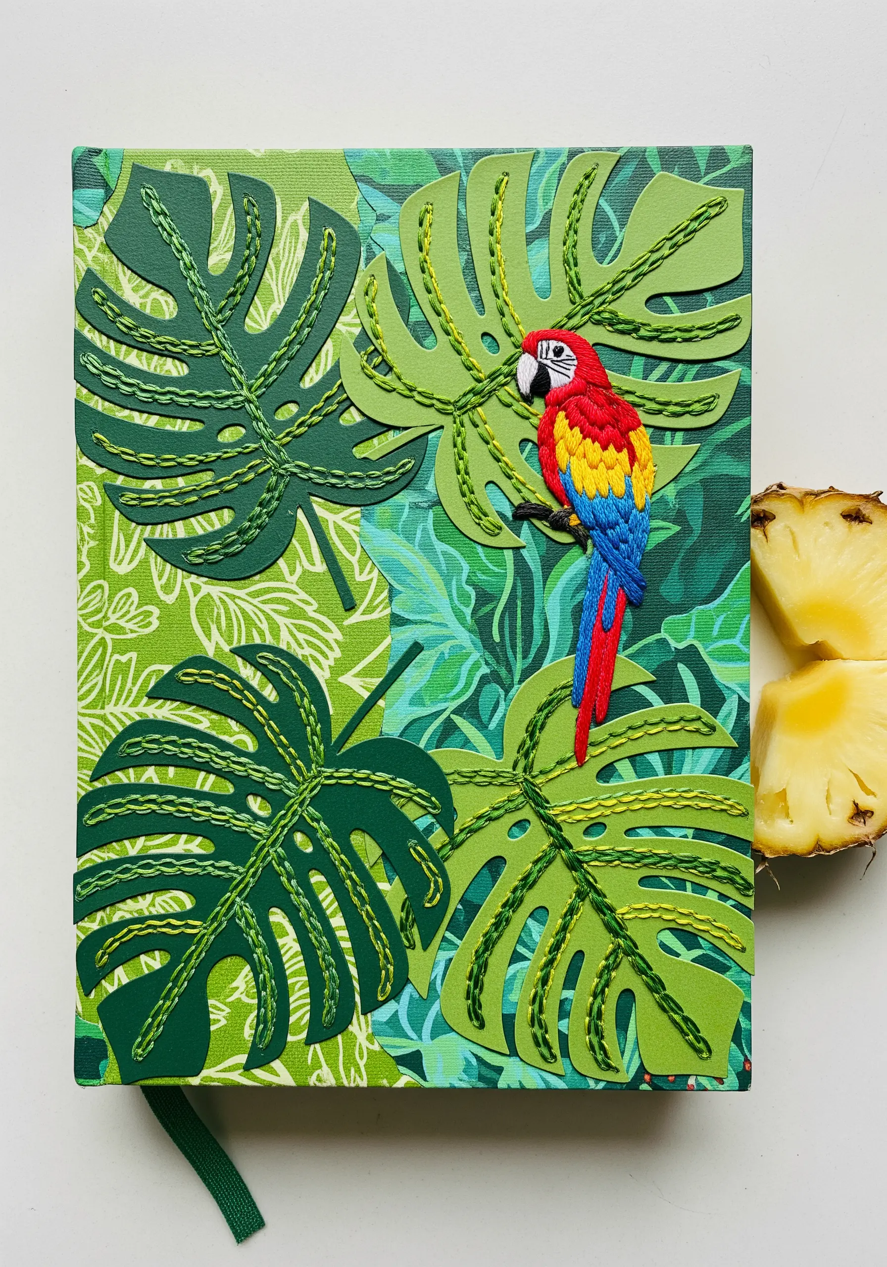 A colorful embroidered parrot perched on layered paper-cut monstera leaves on a journal cover.