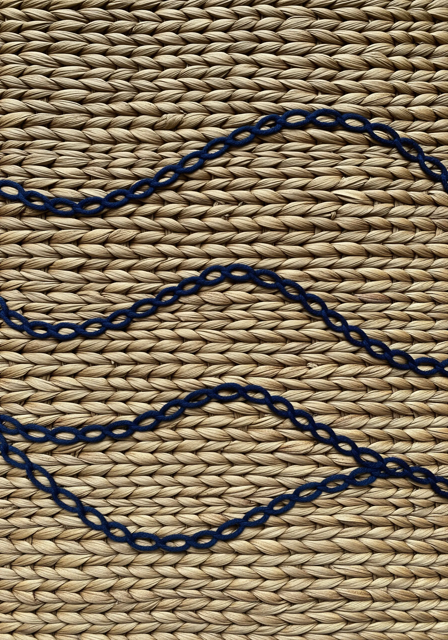 A close-up of a dark blue embroidered chain stitch on a textured, woven seagrass surface.